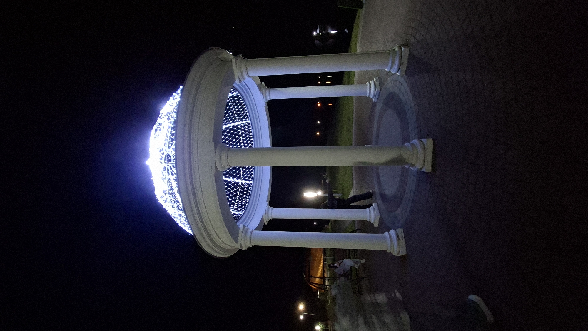 A white, domed gazebo adorned with white lights stands in the foreground at night, its columns illuminated. The gazebo is situated on a brick-paved circular area with a darker circular inlay. To the left of the gazebo, two people are visible, one sitting on a bench and another standing. In the background, a body of water reflects the lights, and a park-like setting with trees and scattered lights is visible. The scene appears to be located in a park in the city of Myadzyel, Belarus.