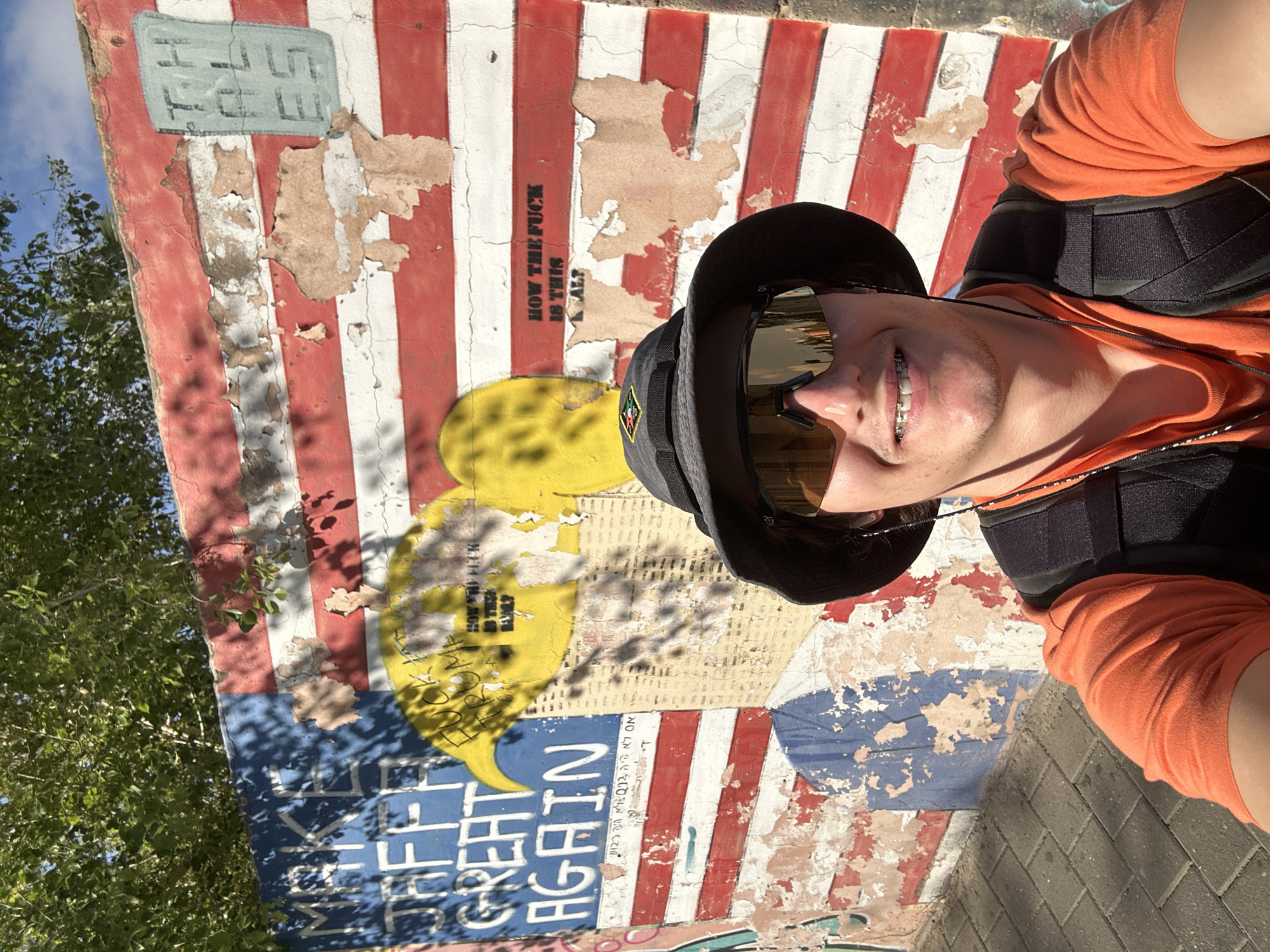 A young man takes a selfie in bright daylight, posing in front of a heavily painted and weathered wall. He is wearing a black bucket hat, mirrored sunglasses, an orange long-sleeved shirt, and a black chest harness or vest. A faint smile is visible, showing dental braces.
The wall behind him features a distressed mural, predominantly red and white stripes, evoking a deconstructed American flag, with a large blue section at the bottom. The paint is peeling extensively, revealing the underlying material. Green foliage and a clear blue sky are visible in the top left, and the ground below is paved with bricks, suggesting an outdoor urban setting in Tel-Aviv, Israel.
Visible graffiti and text on the wall include:
- "HOW THE FUCK IS THIS?" near the upper right.
- Large white letters spelling "GREAT AGAIN" vertically within the blue section at the bottom.
- Hebrew text that reads "אם לא היה Q12 שלא היה 123" (If there wasn't Q12 that wasn't 123) also within the blue section.
- A light blue rectangle at the top left containing some faint white letters that appear to be "TL FE SE".
- Yellow speech bubble shapes are painted on the wall, with some containing additional text like "HOW THE FUCK".
The image captures a casual moment with a backdrop of vibrant, yet decaying, street art under sunny conditions.