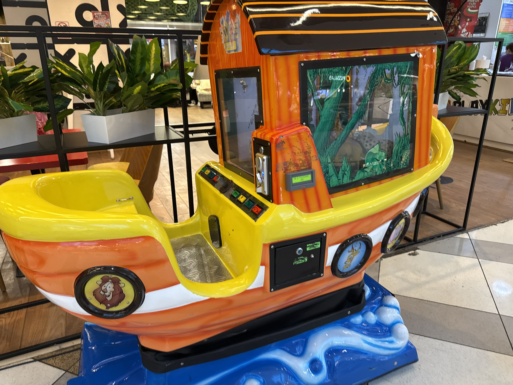 An indoor scene displays a vibrant coin-operated kiddie ride shaped like a boat, positioned on a tiled floor. The boat features an orange hull with a white stripe and yellow trim, resting on a blue base sculpted to resemble water with white waves. A cartoon lion's head is depicted in a circular porthole-like decal on the left side of the hull, and a cartoon monkey is visible in a similar decal on the right. The ride has a yellow cockpit area with a metallic textured floor plate and a control panel featuring red, green, yellow, and blue buttons. A black panel with a keyhole is located on the right side of the hull. A coin mechanism with a blue light and a small digital display showing green text is present on the cabin structure, which has a black roof with orange stripes. Two transparent windows on the cabin display a green jungle-themed background. Text, including "GAJASSIJ" (likely "JASSIG" or "ISSAJAG" reversed) and "PIZZARIA" or "PIZZAS" (reversed), is visible through the window reflections.
In the background, a black metal shelving unit holds several potted plants with green and yellow variegated leaves in white rectangular planters. The flooring is composed of light-colored tiles with darker diagonal lines. Partially visible elements in the background include a red table leg, yellow seating, wooden furniture, and blurred figures of people. Signs are visible on the far walls, including a large black "C" or "G" shape, a red and white sign, and a blue sign with white text resembling "PLAYKIDS". Green-tinted light fixtures are discernible on the ceiling. The setting appears to be a commercial establishment such as a mall or entertainment center in the city of Unknown, Unknown.