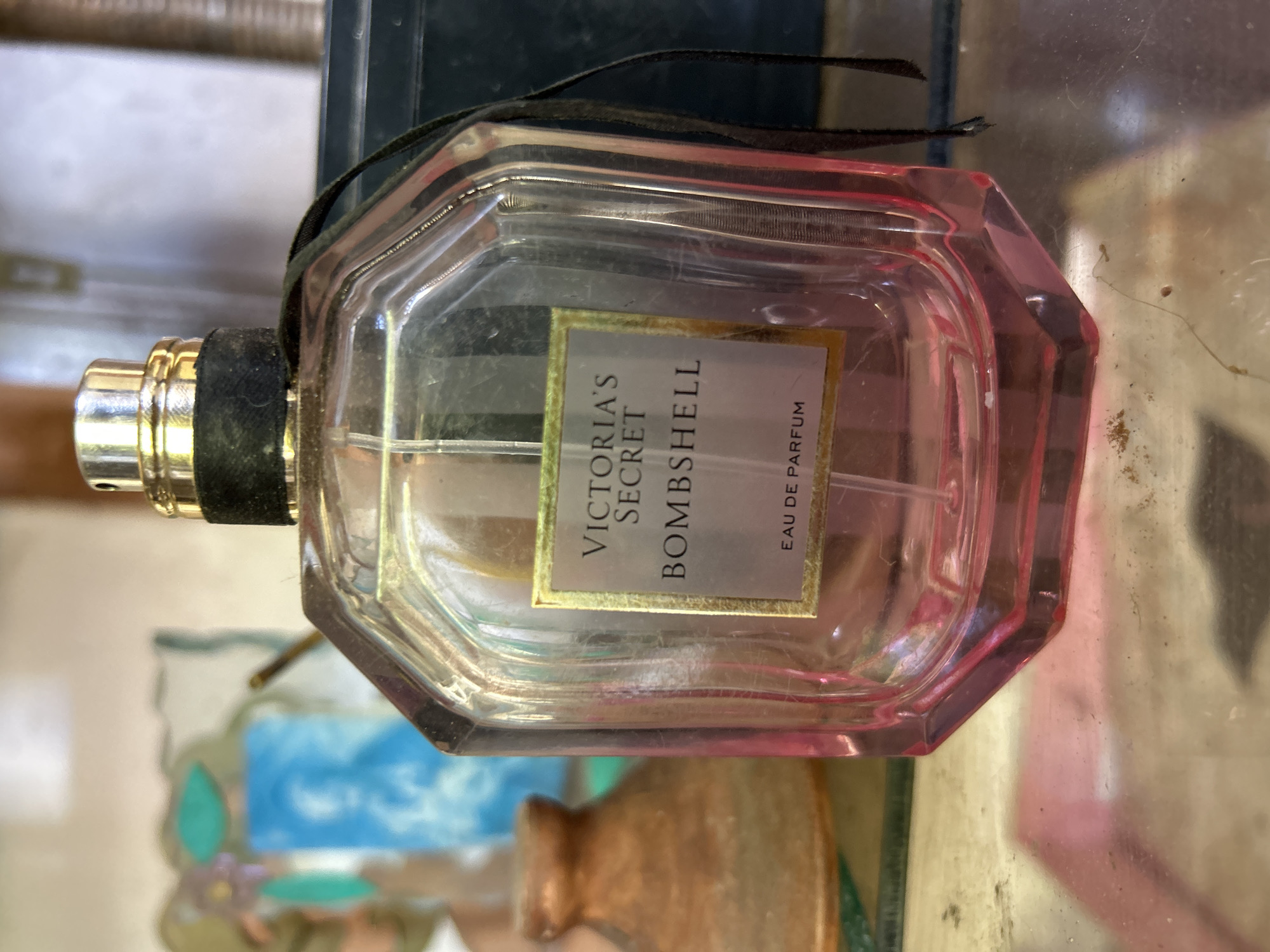 A Victoria's Secret Bombshell Eau de Parfum bottle is the central focus of this image, positioned on a surface that appears to be a shelf or table. The bottle is octagonal in shape with faceted glass, transitioning from a clear top to a pink gradient towards the base. A gold-colored cap with a black band sits atop the atomizer. A thin black ribbon is tied around the neck of the bottle. The label on the front of the bottle is gold with black text that reads "VICTORIA'S SECRET BOMBSHELL EAU DE PARFUM". The background is blurred but suggests an indoor setting. To the left of the perfume bottle, a small, copper-colored, vase-like object is partially visible. To the right, a dark, rectangular object is out of focus. The surface the objects are resting on is cluttered with dust and debris. This scene does not provide any information about a specific location in Santa Ana, Venezuela.
