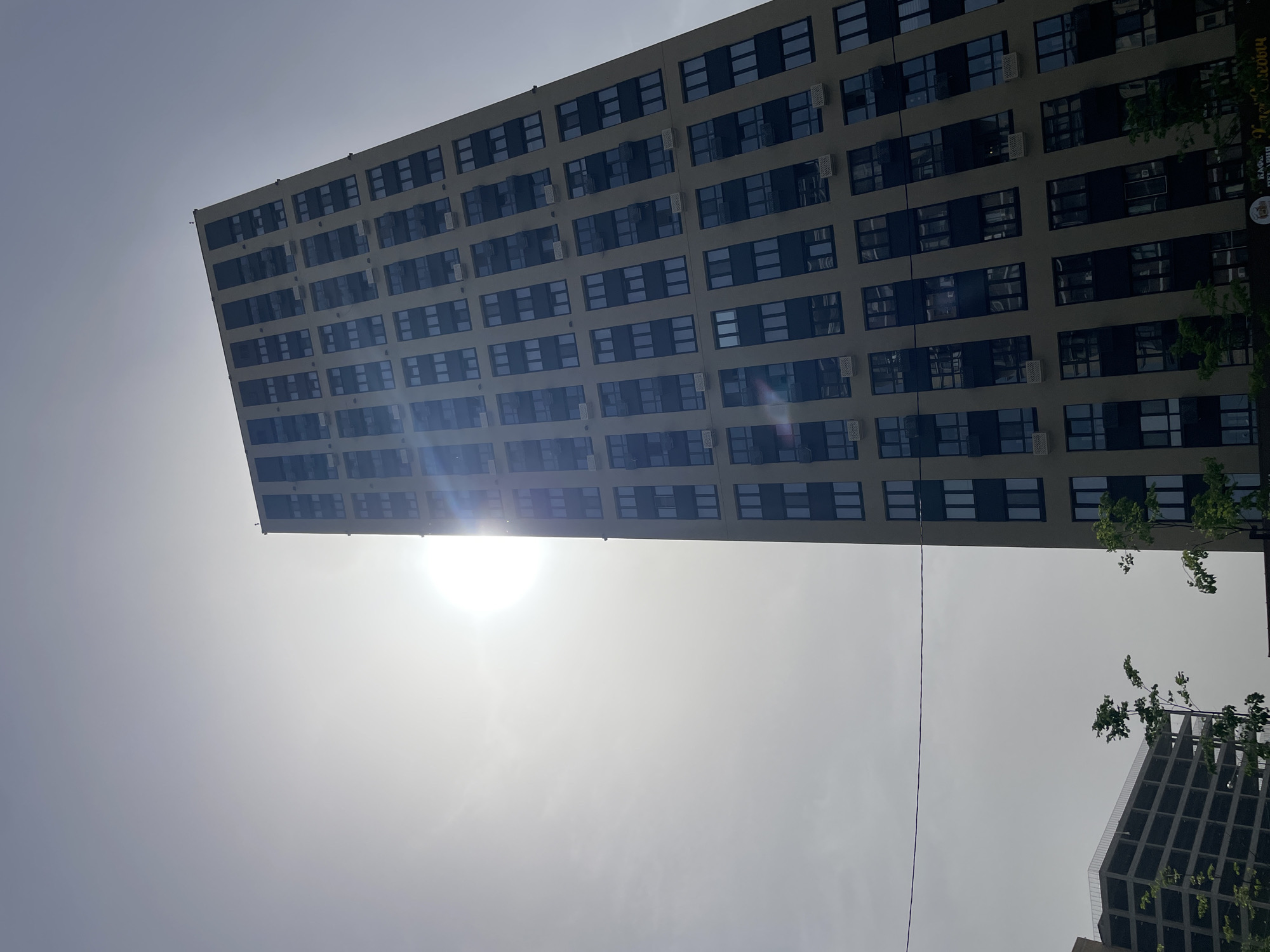 The image shows a tall, rectangular building with many windows under a bright sky, presumably the sun is near. The building appears to be multi-storied and has a somewhat uniform design with rows of windows. In the lower part of the image, the sun is visible. Another building with a similar design is partly visible in the right bottom corner. Based on the sign on the building, this is in Kazan, Russia.