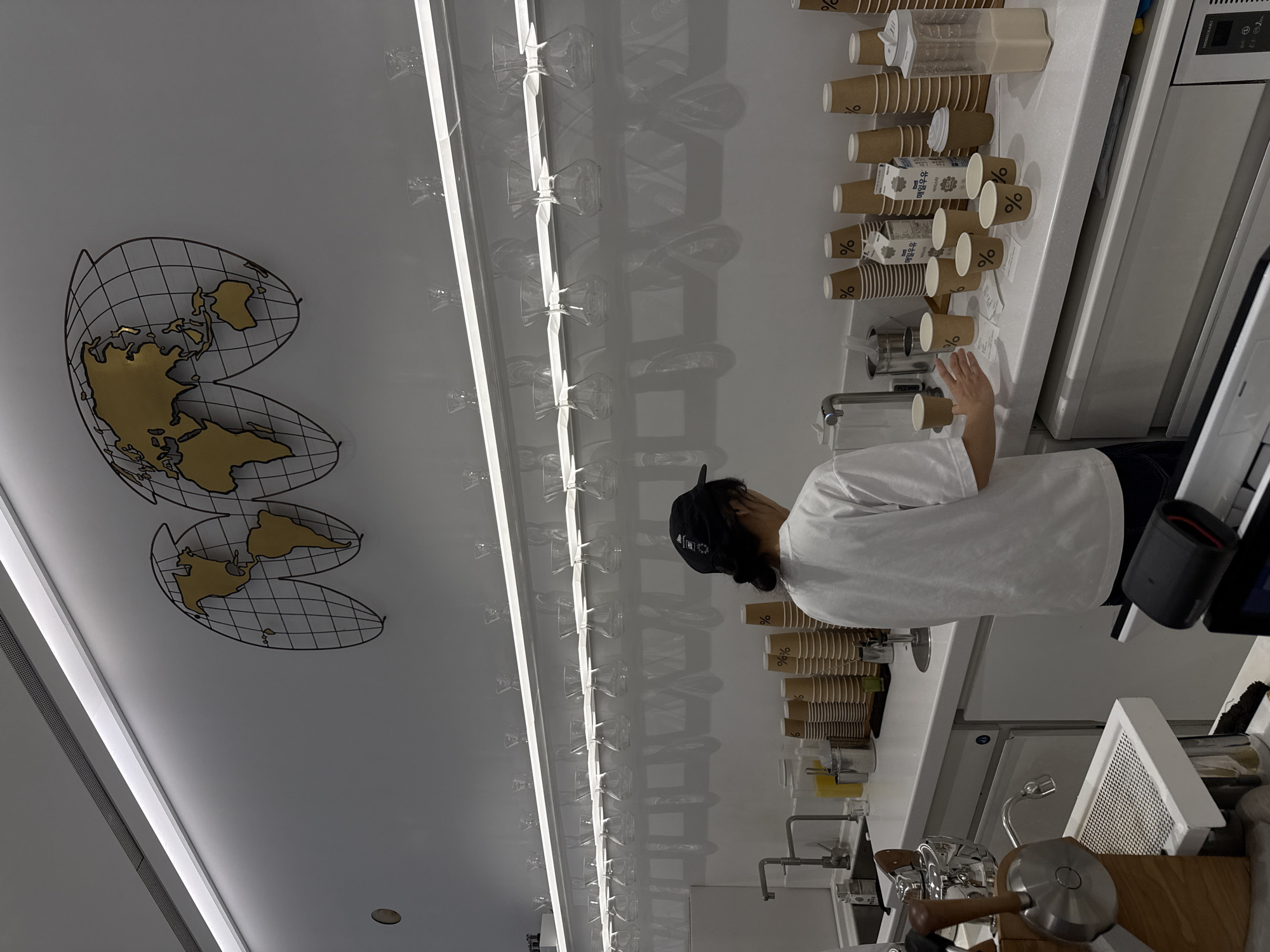 Here is a detailed description of the image:
**Scene:** The image captures the interior of a minimalist-style coffee shop in Seoul, South Korea, likely part of the "Arabica" coffee chain.
**Environment:** The shop features a clean, white aesthetic with multiple rows of glasses hanging above the counter. A wireframe world map adorns the wall.
**People:** A person wearing a white t-shirt and a black baseball cap is visible. They are standing at the counter.
**Objects:** Coffee cups with the "%" logo of the Arabica coffee brand are stacked on the counter. There's coffee brewing equipment, including espresso machines and sinks with faucets. A refrigerator or cooler is also visible. Milk cartons are placed next to the coffee cups.
**Actions:** The person is likely interacting with the coffee-making equipment, potentially preparing a beverage.
**Interaction:** The person appears to be working at the coffee shop.