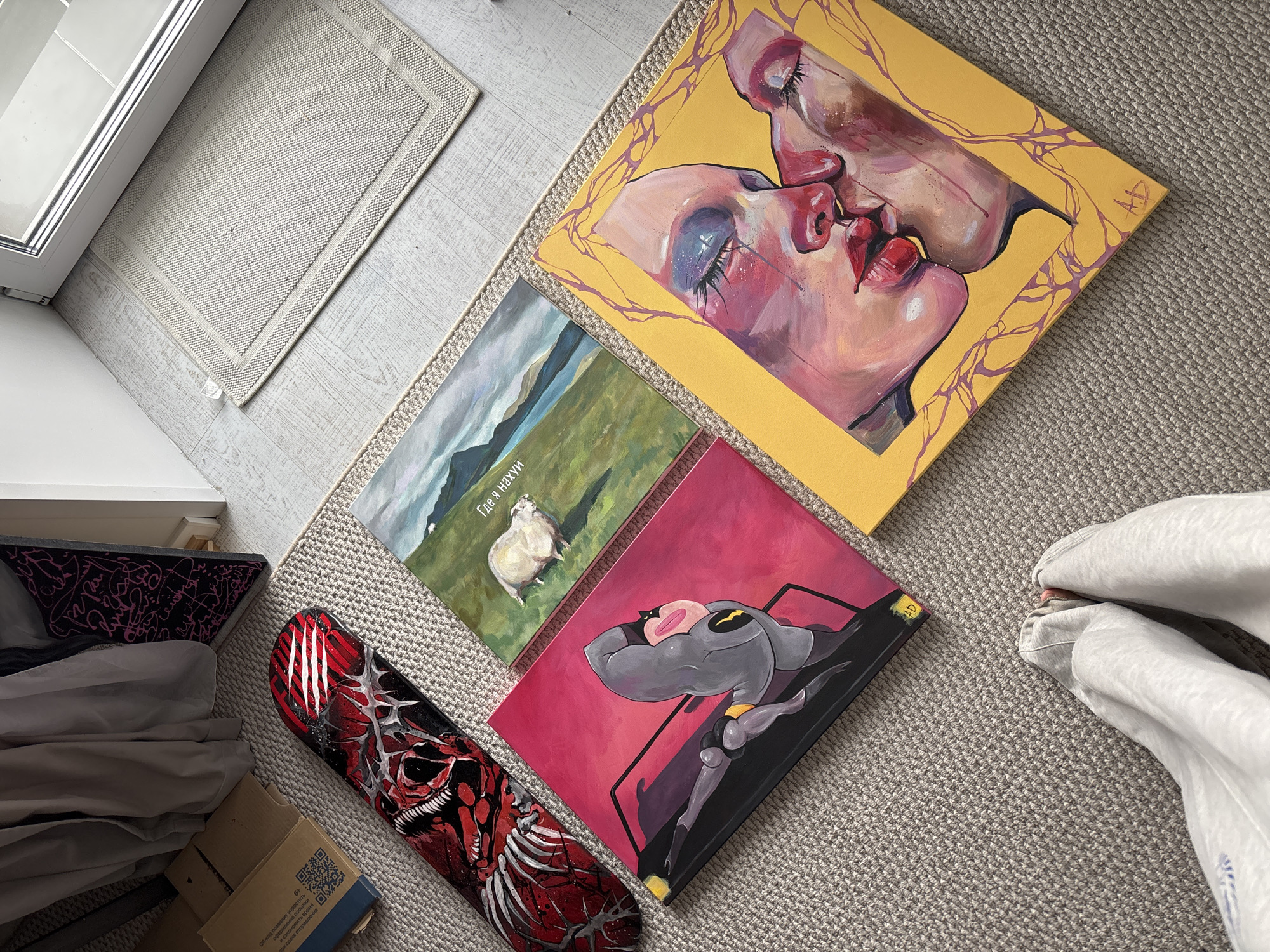 The image is an eye-level, medium shot of several objects and art pieces laid out on a beige carpet. In the top left corner, there is a white doormat adjacent to a white door frame. To the left of the doormat, there is a stack of items, including a piece of black and pink art, folded white fabric, and a cardboard box with Russian text and a QR code, suggesting possible origin in Krasnoyarsk, Russia.
In the upper right, there is a square canvas art piece depicting two people kissing against a yellow background. To the lower-left of this painting, there is another square canvas with a green landscape featuring a sheep and Russian text reading "Где я нахуй". Below this, there is another square canvas with an illustration of a cartoon character against a pink background.
To the left of the cartoon canvas, there is a snowboard with a red and silver skeletal design. In the lower right corner, the bottom half of a person wearing grey sweatpants is visible. The entire scene is lit by natural light.