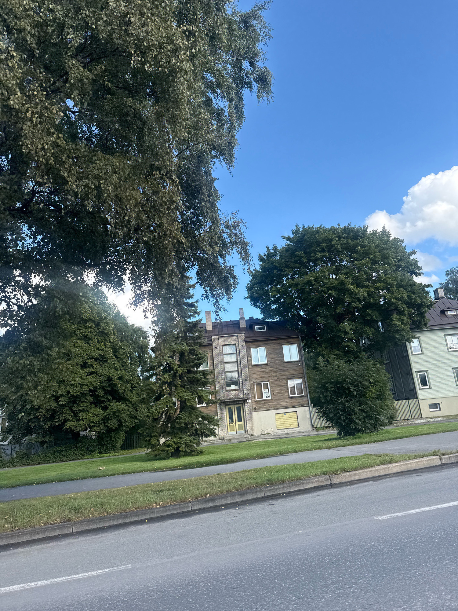 In Tallinn, Estonia, a wide asphalt road runs horizontally across the foreground, with a white line painted on its surface. Beyond the road, a strip of manicured green grass runs along a curb, separating the road from a sidewalk. Behind the sidewalk, more green grass and trees form a natural barrier. In the background, several buildings are visible. To the left, a building with grey brick and dark trim has multiple windows and a prominent chimney. Next to it is a building with dark brown wooden siding, featuring several windows and a yellow door. Further to the right, a light green building with dark trim and windows is situated. Large, mature trees with dense green foliage dominate the landscape, with one particularly large tree in the center partially obscuring the buildings. The sky above is a clear blue with scattered white clouds.
