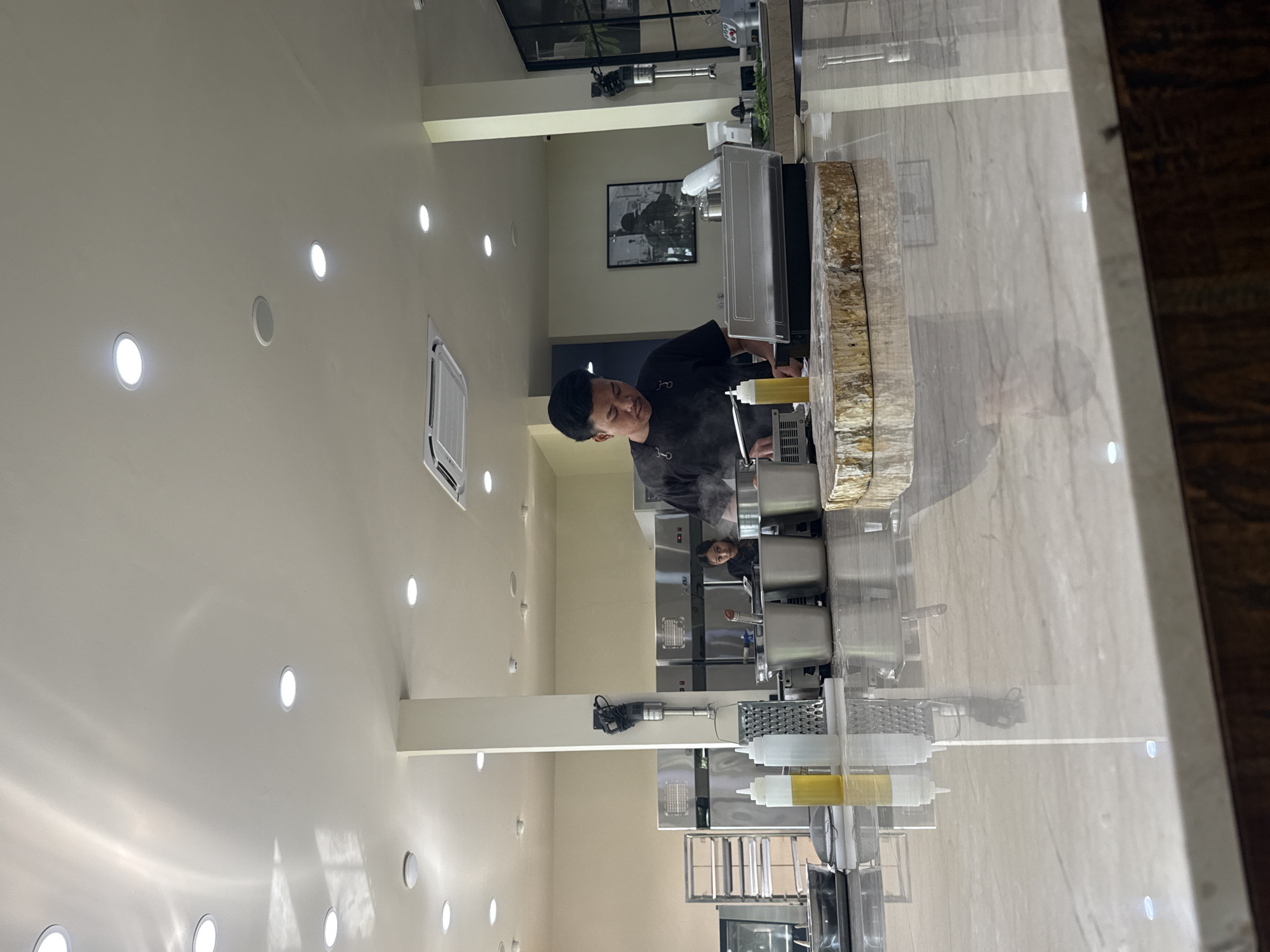 In Mengwi, Indonesia, a chef is preparing food behind a counter. The counter has a decorative, natural-edged design. Behind the chef is a stainless steel food preparation area with various containers and equipment. Other staff are visible further back. The ceiling is white with multiple recessed lights. A framed picture hangs on the wall behind the preparation area.