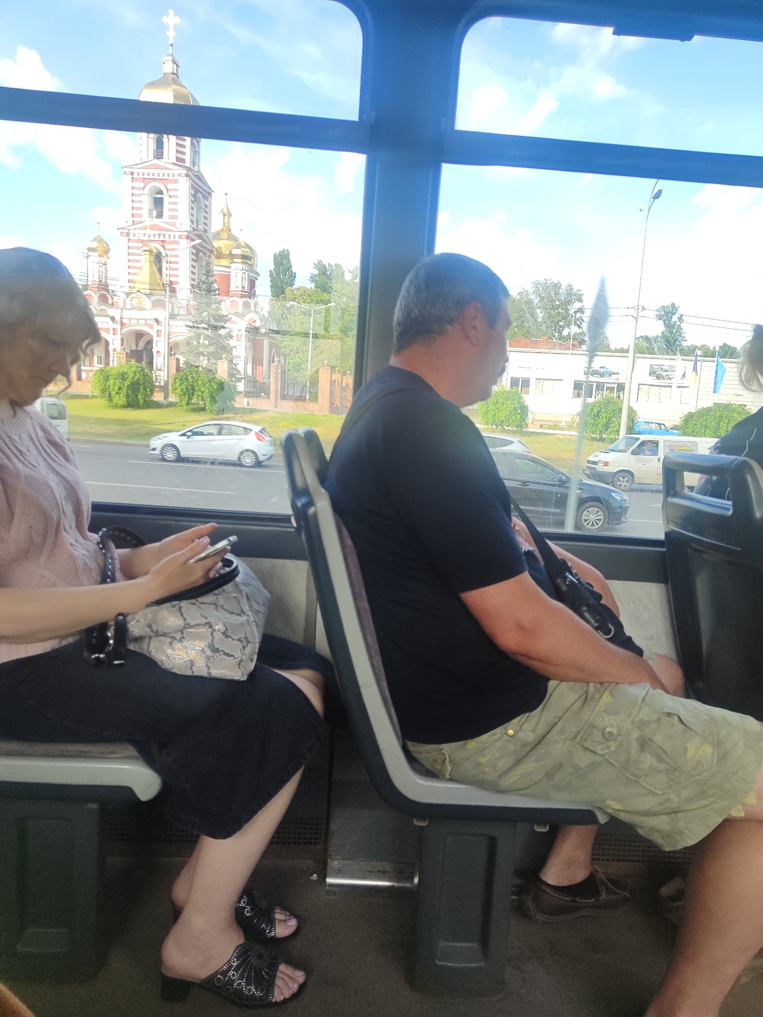 This is a medium shot photograph taken from inside a vehicle, likely a bus or train, looking out a window. The scene is bright, suggesting daylight, possibly a sunny day given the clear sky and the light illuminating the church.
The primary subjects are two individuals seated on the right side of the frame. On the left, a woman with blonde hair is looking down at her phone, which she holds in her hands. She is wearing a light pink knitted top and a black skirt. A patterned purse rests on her lap. On the right, a man with short gray hair is seen from the side, looking forward. He is wearing a black t-shirt and light green camouflage-patterned shorts. A dark bag is slung across his body.
Outside the window, a white car is visible on a road. In the background, a large, ornate church with red and white striped walls and golden domes dominates the scenery. Trees and other buildings are also present. The sky is blue with some scattered white clouds. The overall mood appears relaxed, with the passengers engaged in their own activities. The presence of the church suggests a location with religious architecture, possibly in a town or city.