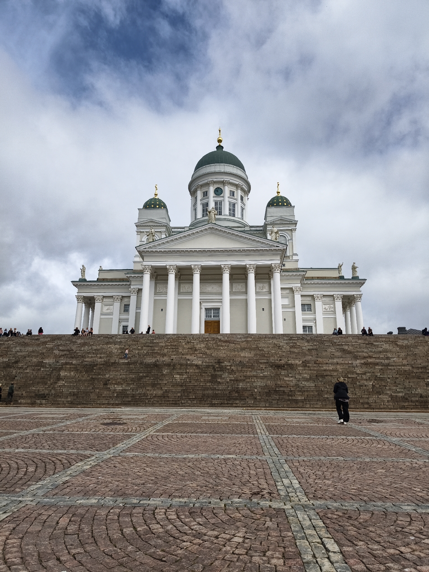 A low-angle, wide shot captures the Helsinki Cathedral, a neoclassical landmark, under a cloudy sky. The cathedral features a large central dome and two smaller domes, all topped with gold crosses. A grand staircase made of stone leads up to the building, which is adorned with white columns and classical statues. A cobblestone plaza with a geometric pattern stretches across the foreground. Several people are scattered throughout the scene, with some sitting on the stairs and one person walking across the plaza. The overall atmosphere is solemn and majestic, with the diffused light from the overcast sky adding a dramatic effect.
