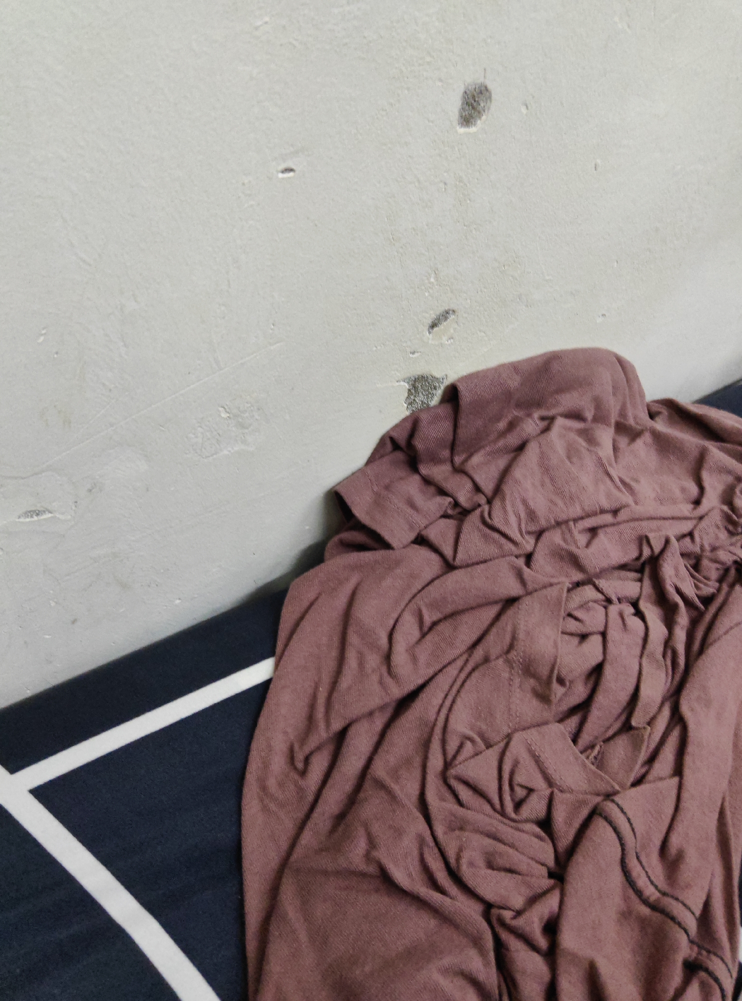 A close-up, high-angle shot captures a crumpled, dusty rose-colored t-shirt resting on a dark blue and white striped surface. The t-shirt is heavily creased, with visible seams and a dark thread along one edge. To the left of the t-shirt, a light gray wall is visible, showing signs of wear with several dark marks and chipped paint. The lighting is even, with no harsh shadows. The scene is static, with no people or animals present. The image appears to be taken indoors, possibly in a residential setting.