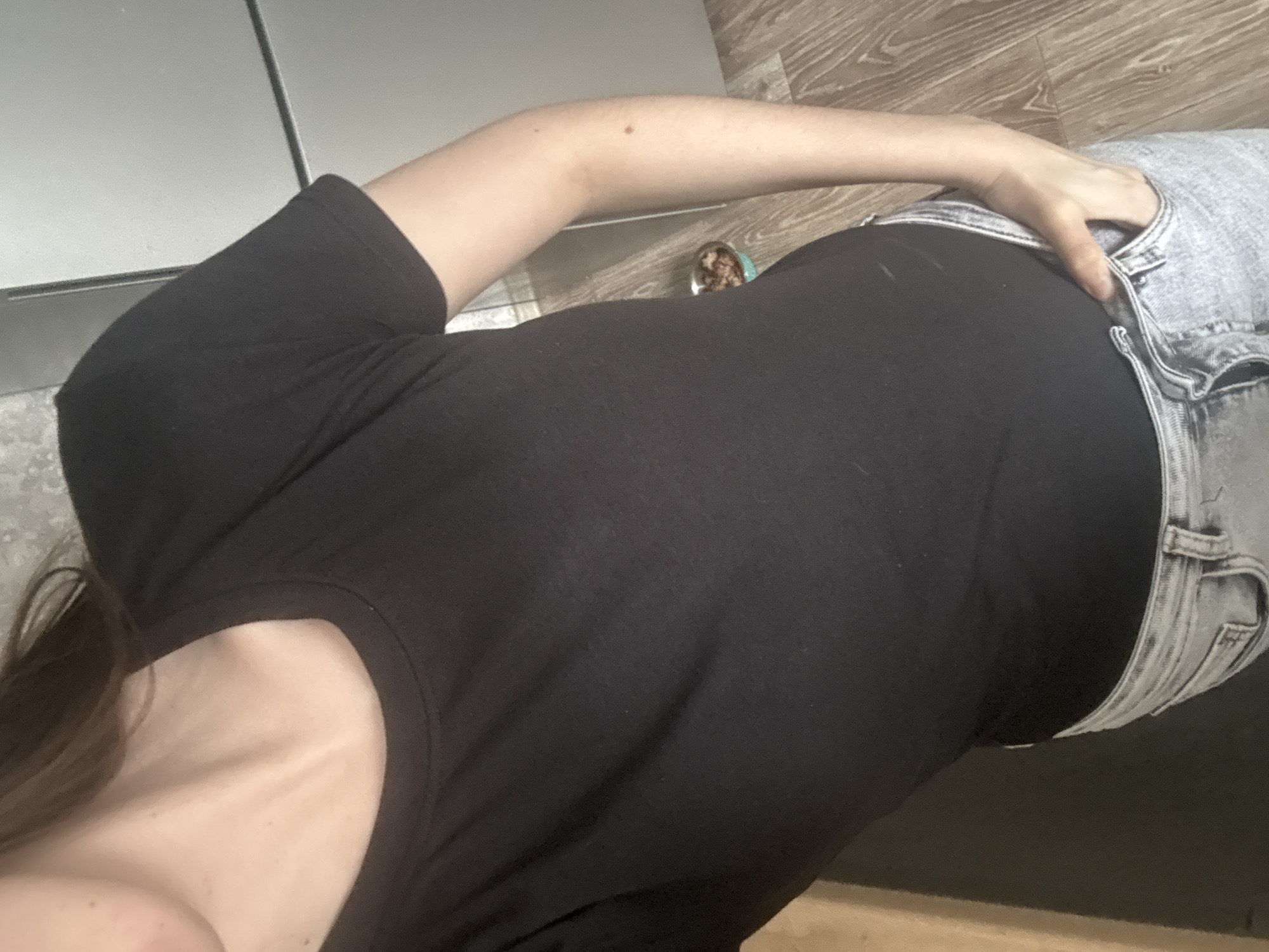 A woman is shown from a high-angle perspective, wearing a black t-shirt and light-wash denim shorts. Her left hand rests on the upper right part of the denim shorts. The setting appears to be indoors, on a wooden floor. The location details of Chaikovsky, Russia, are not evident in the image.