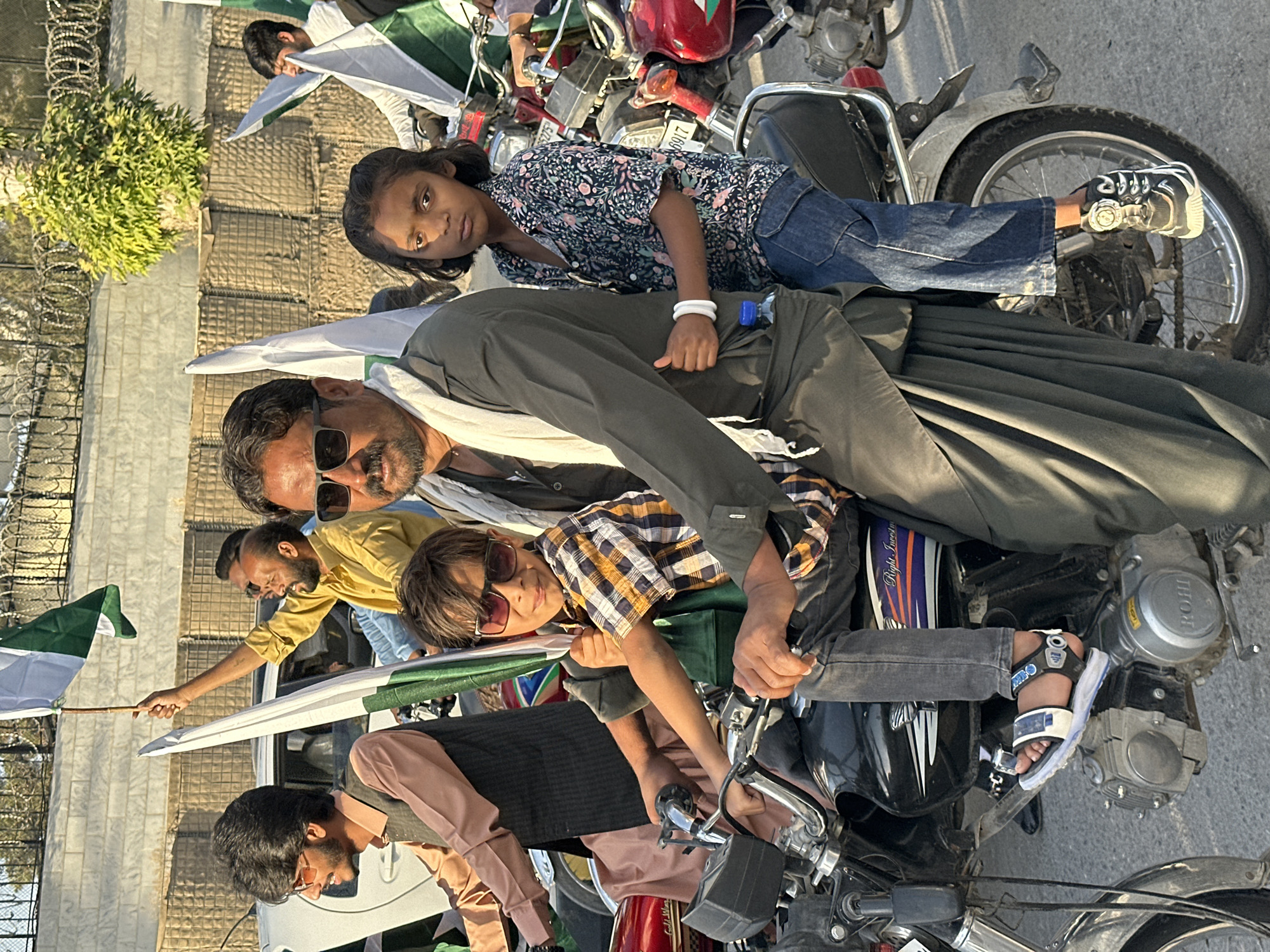 A group of people, including adults and children, are densely packed on a collection of motorcycles in an outdoor setting. Several individuals hold Pakistani flags. The scene appears to be a demonstration or celebration. The background includes a structure with a tall wall topped with barbed wire. The setting is likely in Quetta City Tehsil, Pakistan.