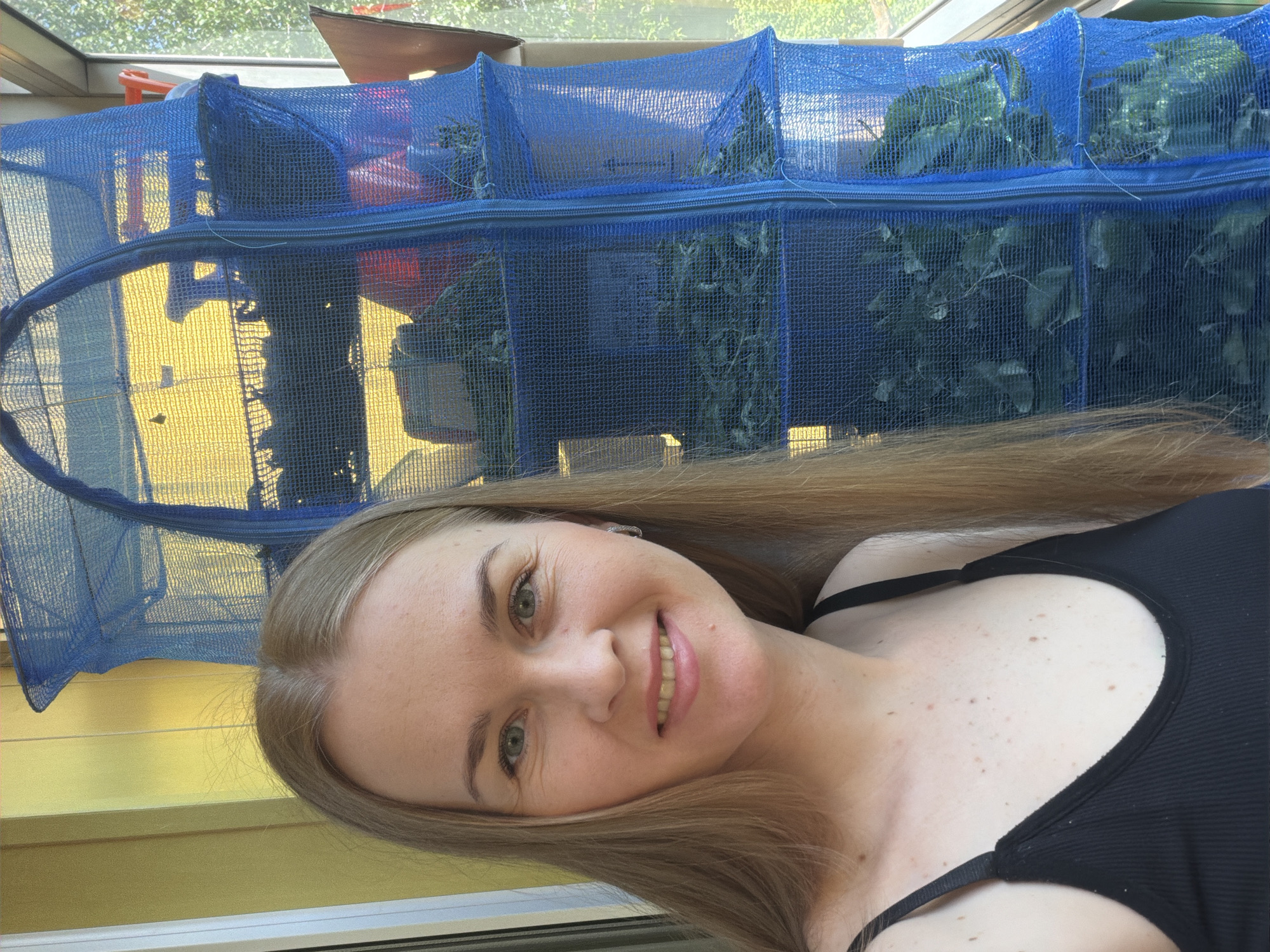A blonde woman with light green eyes and fair skin is taking a selfie on a balcony. She is wearing a black tank top. Behind her, to the right, is a blue mesh drying rack with multiple shelves, filled with what appear to be dried leaves or herbs. The balcony overlooks a yellow wall and a green, leafy exterior. The lighting suggests it is daytime. The background details are consistent with an urban environment in Khabarovsk, Russia.