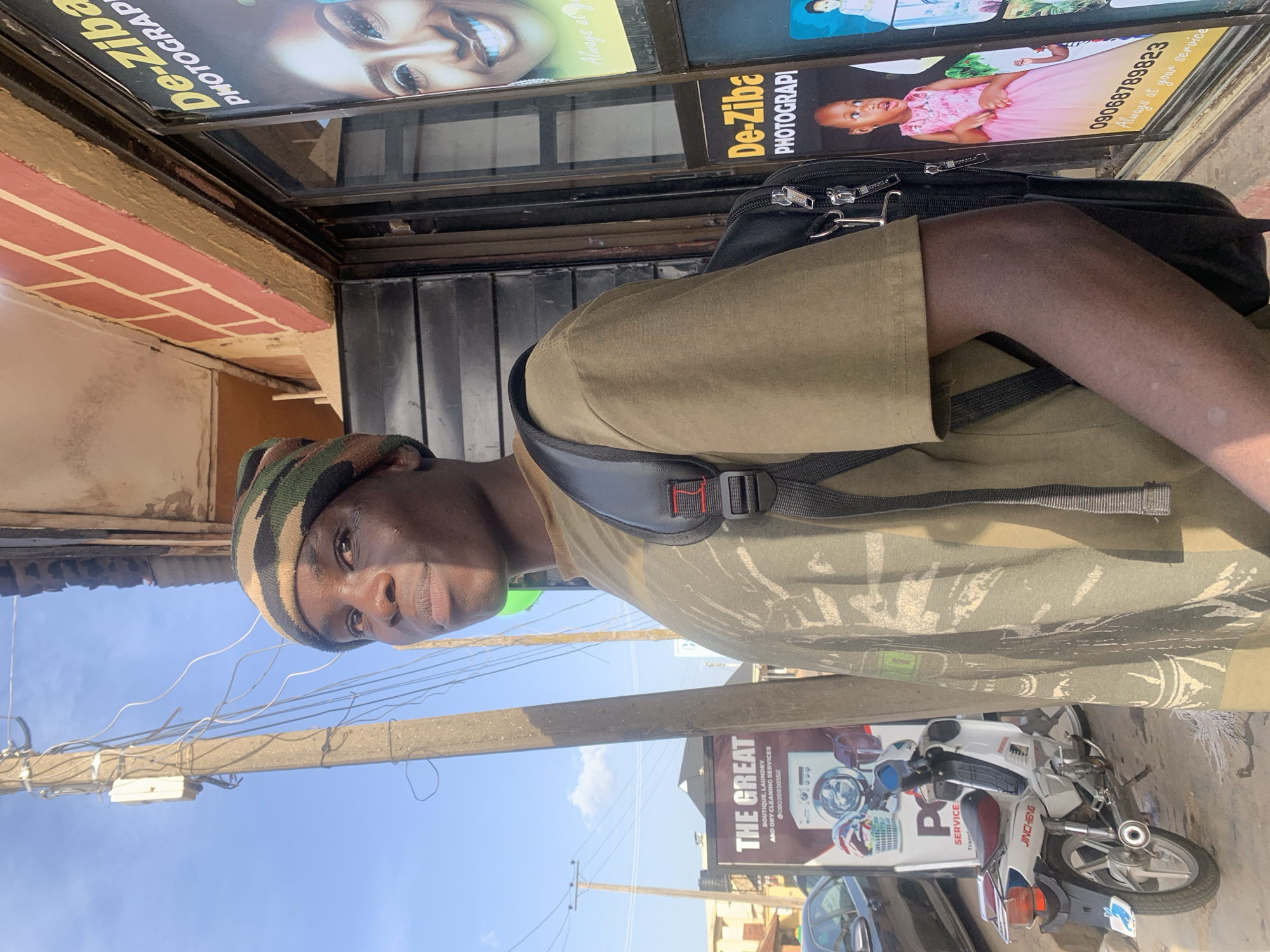 A street-level shot in Jos, Nigeria. An individual with a striped beanie is wearing a green t-shirt with a black backpack, facing the camera. The background includes a white and red motorcycle parked near a sign advertising "The Great Boutique, Laundry, and Dry Cleaning Services" and a photography business named "De-Ziba Photography." There are also parked cars, utility poles with wires, and building facades visible. The sky is clear and blue.