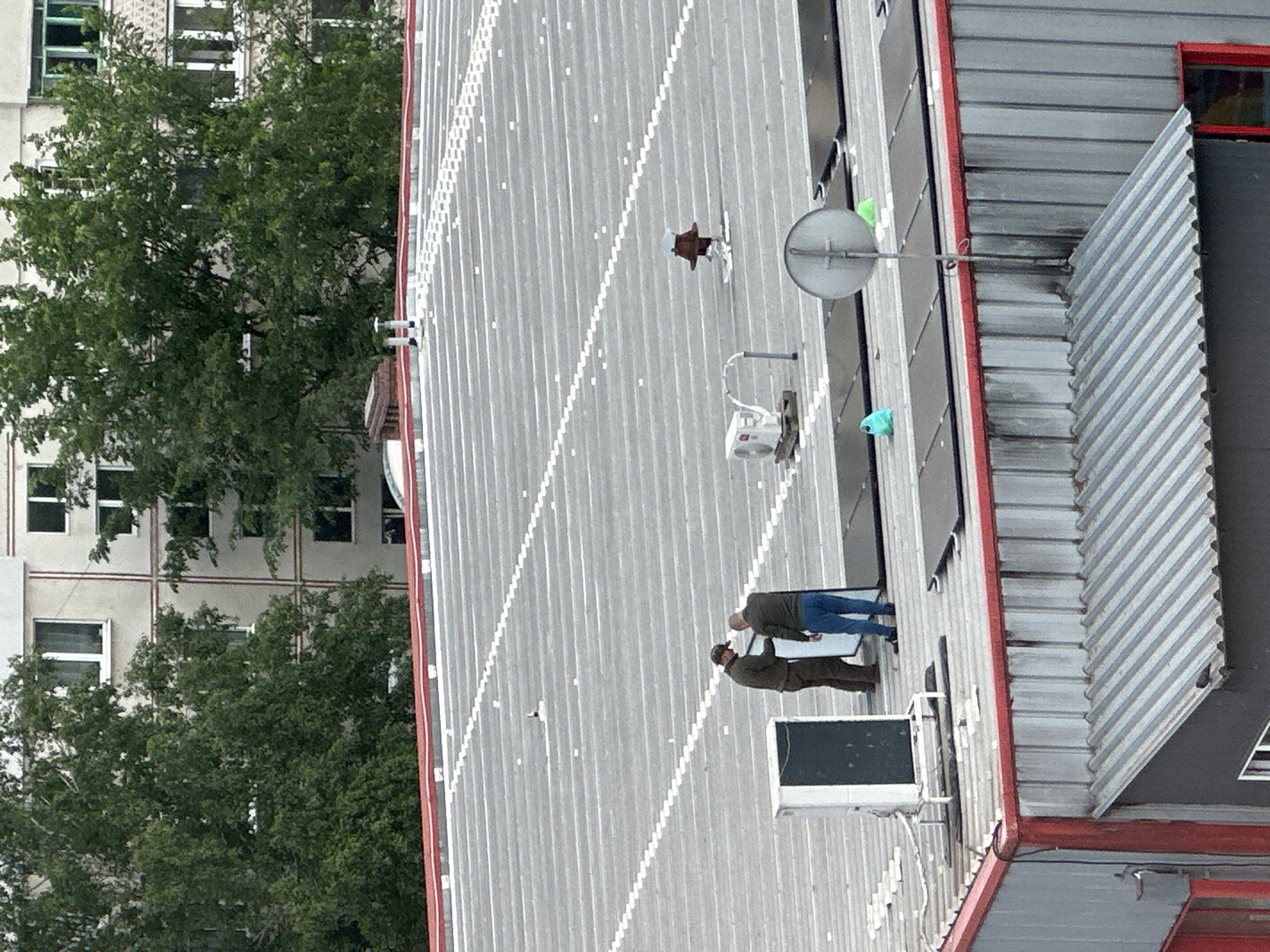 The scene appears to be a rooftop in an urban environment. The rooftop is made of corrugated metal with what seem to be solar panels installed. Two men are standing on the roof; one is wearing a dark green jacket and khaki pants, while the other wears a grey top and blue jeans. Nearby is a satellite dish, an air conditioning unit, and several unknown objects. In the background, there's a building with multiple windows obscured by dense green trees. The location is Unknown, Unknown.