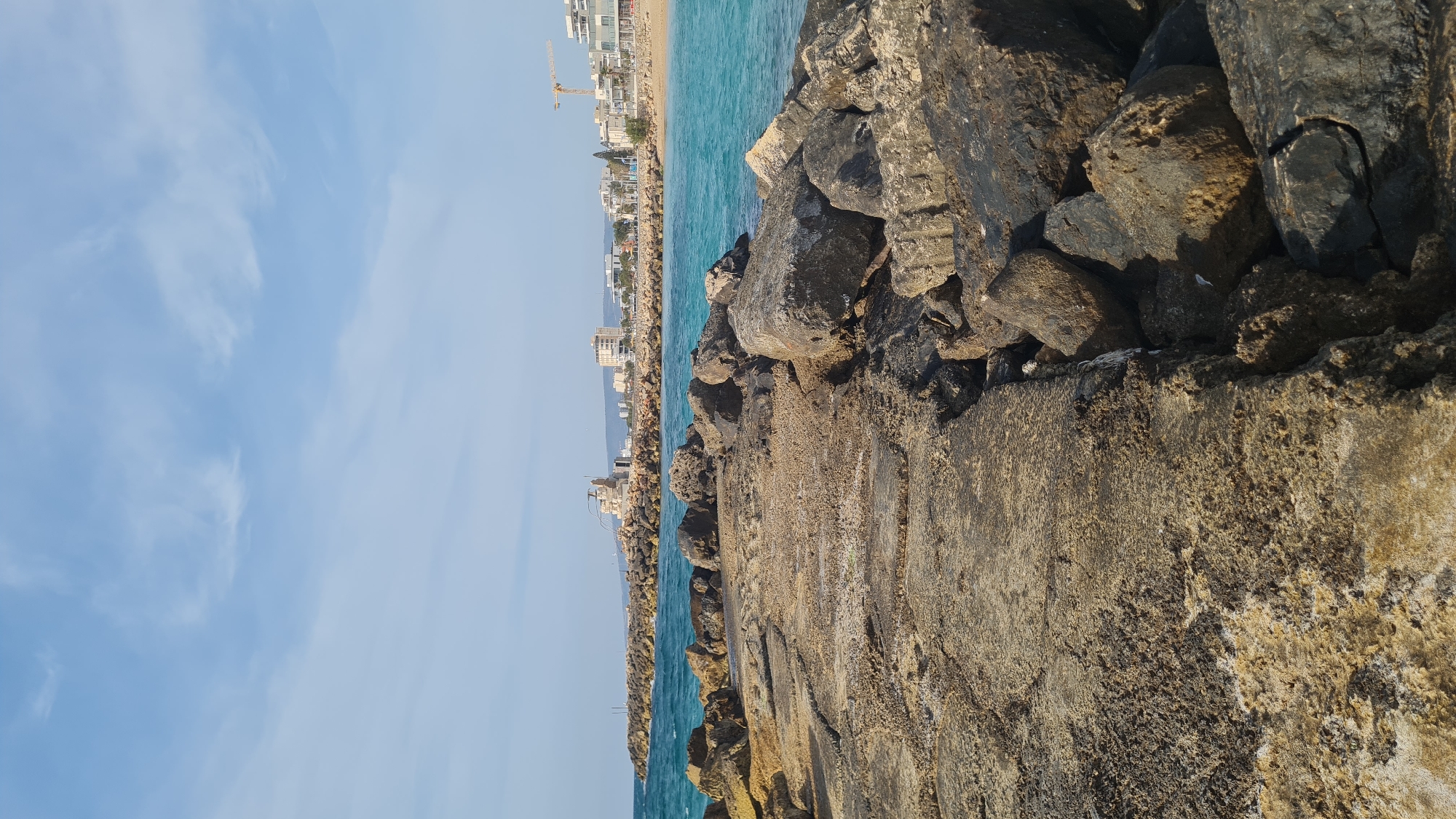 The image depicts a bright, clear day along a coastal area, viewed from a rocky breakwater. In the immediate foreground, a rough, textured concrete or stone surface extends, with dark, irregularly shaped rocks piled on its right side, forming part of the breakwater. These rocks show varying shades of grey and brown, with some appearing wet or darkened by moisture.
To the right of the breakwater, the calm, turquoise-blue ocean stretches into the distance, meeting a sandy shoreline. Along this distant shore, a city skyline is visible, featuring numerous modern buildings, some appearing residential or commercial, under a bright sun. A tall yellow construction crane stands out against the sky, indicating ongoing development in the area. Another section of the rocky breakwater or coastline can be seen further along the sea, leading towards the city.
The sky, occupying the upper left portion of the image, is a vibrant clear blue with soft, wispy white clouds scattered across it. The lighting suggests it is daytime, likely late morning or early afternoon, with sunny and fair weather conditions. There are no people visible, nor any specific activities taking place. No discernible text is present in the image.