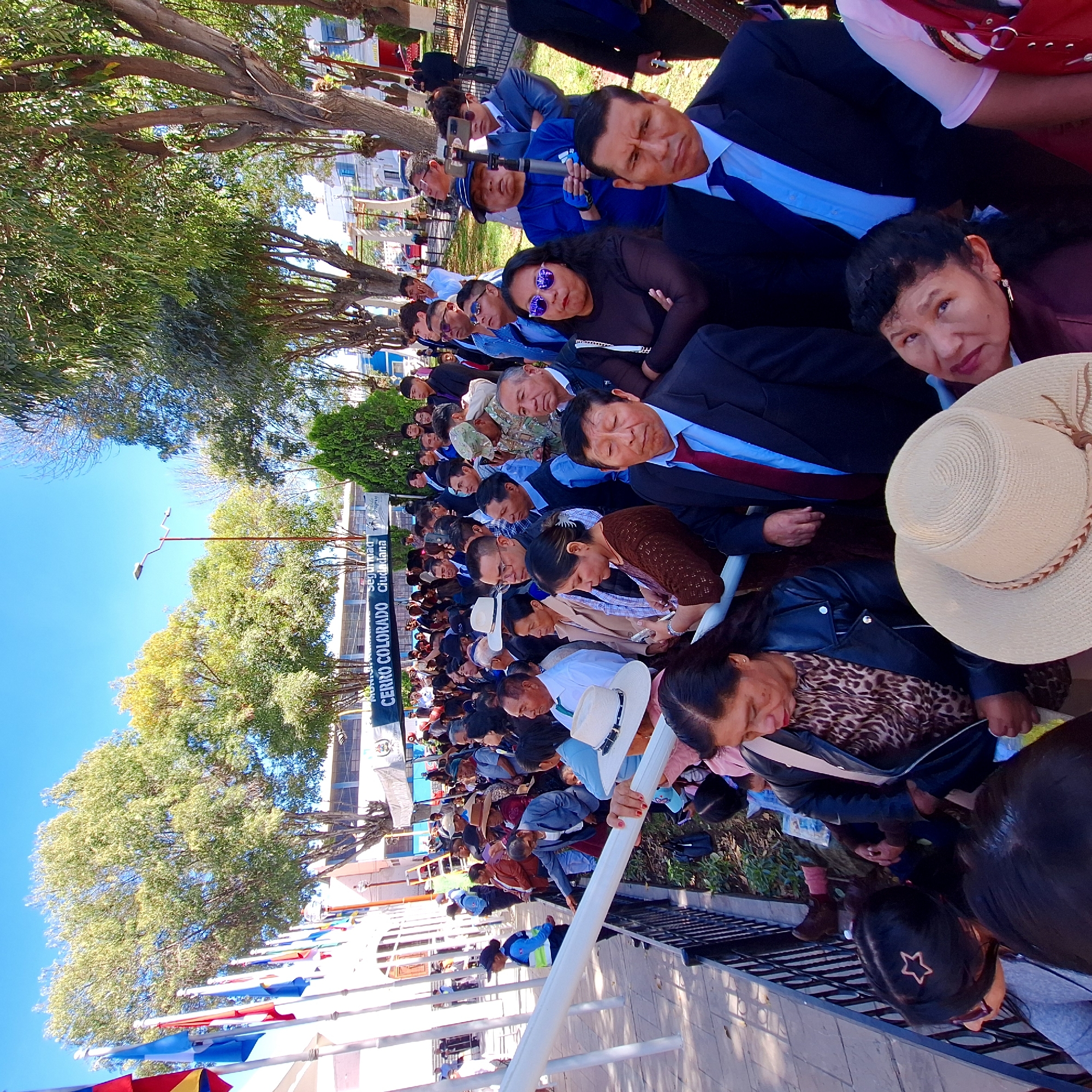 A large group of people, dressed in a mix of casual and formal attire, are gathered outdoors in what appears to be a public space. The banner of Cerro Colorado is visible. Most of the people are seated and facing in the same direction, suggesting they are watching an event or presentation. Trees provide shade over the scene. Several individuals are wearing hats, and some are wearing sunglasses. The location is Cerro Colorado, Peru.