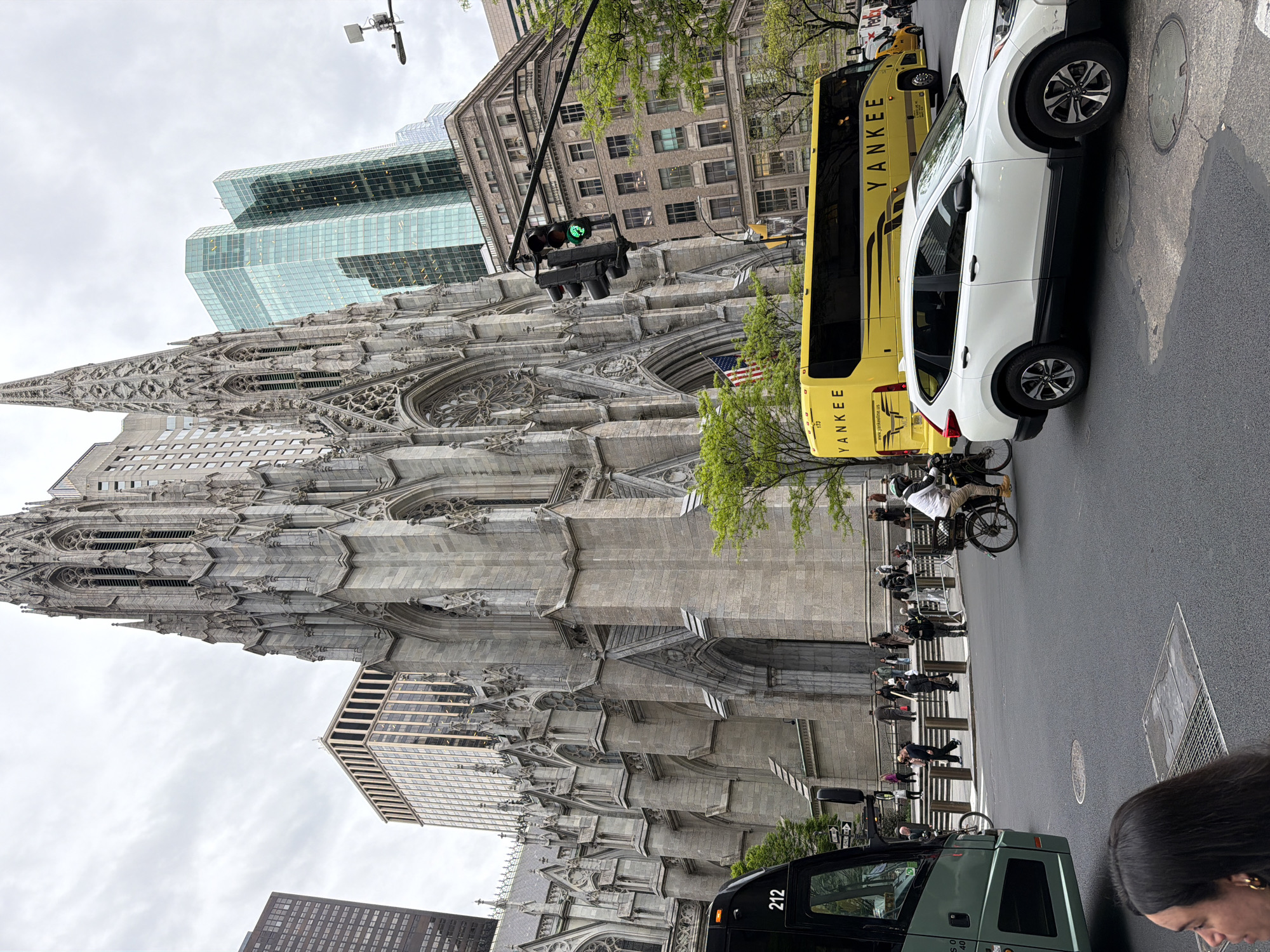This image, rotated 90 degrees clockwise, captures a dynamic street scene in New York City, dominated by a grand Gothic Revival cathedral. The main subject is St. Patrick's Cathedral, with its intricate stone carvings, towering spires, rose window, and visible flying buttresses, showcasing elaborate architectural details. An American flag is displayed near one of its entrances.
In contrast to the historic cathedral, several modern and older buildings frame the background, including a prominent glass skyscraper with a distinctive layered design and an older brick building.
At street level, the scene is bustling with activity. A bright yellow tour bus, clearly marked "YANKEE" on its side and with the number "173", is parked or moving on the right side of the street. Next to it is a white SUV. Further down the street, a dark green bus with the number "212" on its front is partially visible in the bottom foreground. A traffic light, showing a green signal, hangs above the street.
People are visible on the sidewalk, including pedestrians walking and a person riding a bicycle or pedicab. In the bottom right corner, the top of a person's head with dark hair is visible, suggesting the photo was taken from a low angle or by someone close to the ground. The weather appears overcast, indicated by the gray sky, suggesting daytime. Manhole covers and street markings are visible on the asphalt road. A "ONE WAY" sign is also partially visible on a pole.