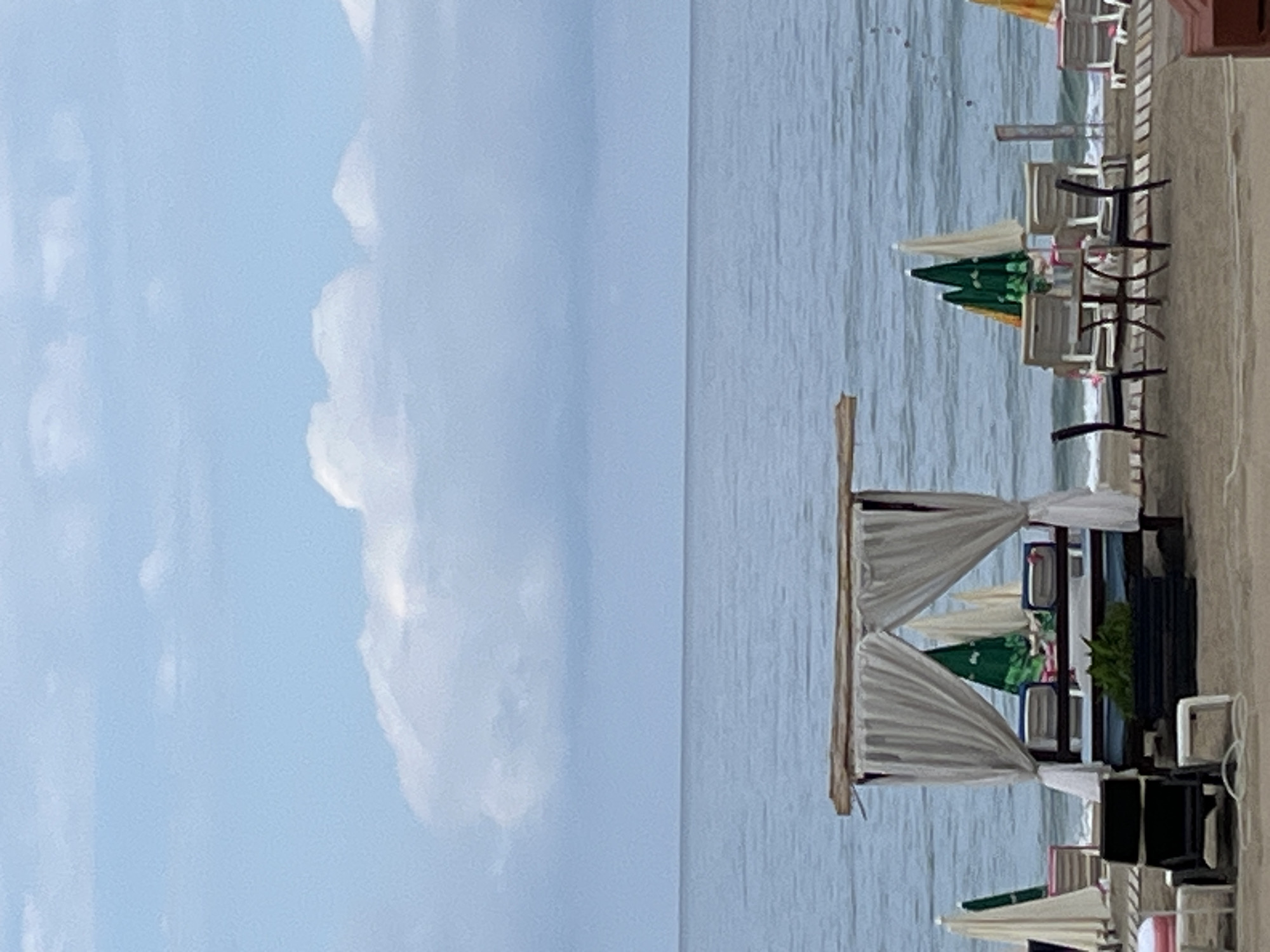 The image depicts a beach scene in Alanya, Turkey. On the right side of the frame, along the shoreline, are several rows of lounge chairs, umbrellas (mostly green and white), and beach cabanas with white fabric drapes. Beyond these, the calm, light-blue water stretches toward the horizon. The left side of the frame is dominated by a light blue sky with fluffy white clouds. The image is rotated 90 degrees counterclockwise.