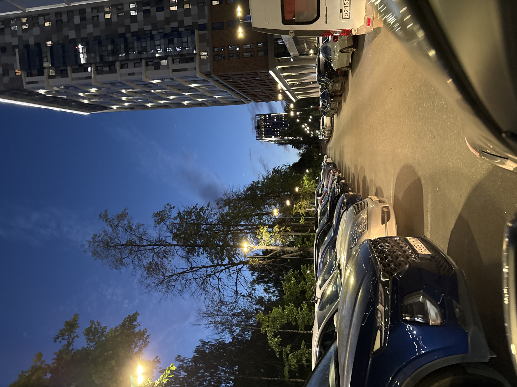 An outdoor, low-angle, medium-full shot captures a parking lot filled with cars at dusk, located in Izhevsk, Russia. On the right, a tall, modern residential building with illuminated windows and a brick facade dominates the scene. To the left, a line of trees with sparse foliage stands tall against the twilight sky, punctuated by streetlights casting a warm glow. The parking lot surface is asphalt, and multiple cars are parked in rows, facing away from the viewer. The foreground shows the hood of a dark blue SUV, its headlights on, reflecting the surrounding lights. In the distance, another taller building with illuminated windows is visible. The sky is a deep blue with wispy clouds.