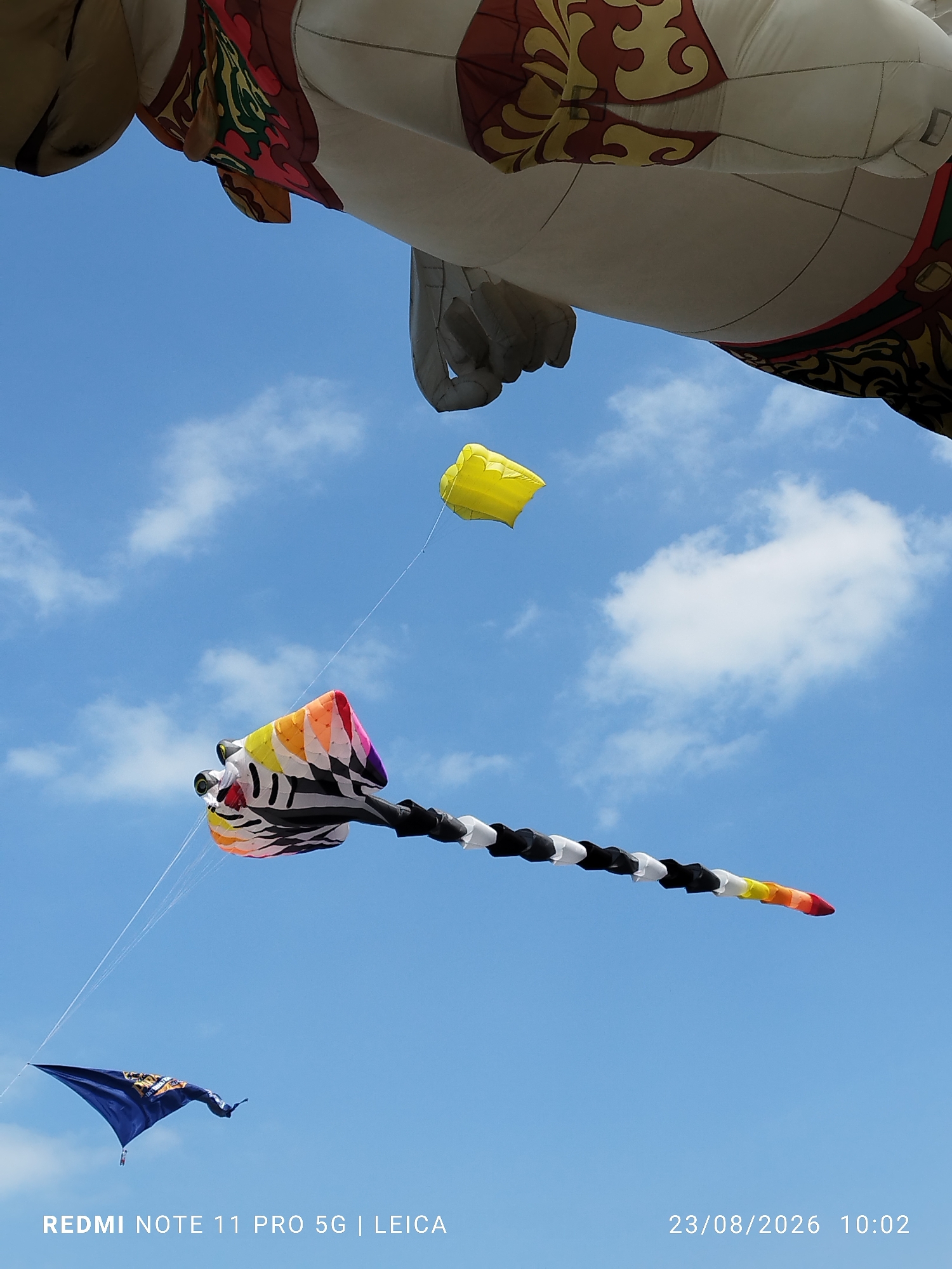A bright blue sky with scattered white clouds serves as the backdrop for an outdoor scene, likely a kite festival, in Parangtritis, Indonesia. In the foreground, a large, white, hot air balloon shaped like a mythical creature, possibly a dragon or horse, with intricate red and yellow decorative patterns, dominates the upper portion of the frame. Below the balloon, a yellow kite is suspended in the air. Further down, a long, colorful kite with a black and white striped tail stretches across the sky, its head featuring a rainbow pattern and a red accent. A small, dark blue kite with an orange and yellow emblem is visible in the lower left corner. The lower right corner displays a timestamp: "23/08/2026 10:02" and text identifying the device used: "REDMI NOTE 11 PRO 5G | LEICA". No people are visible in the image.