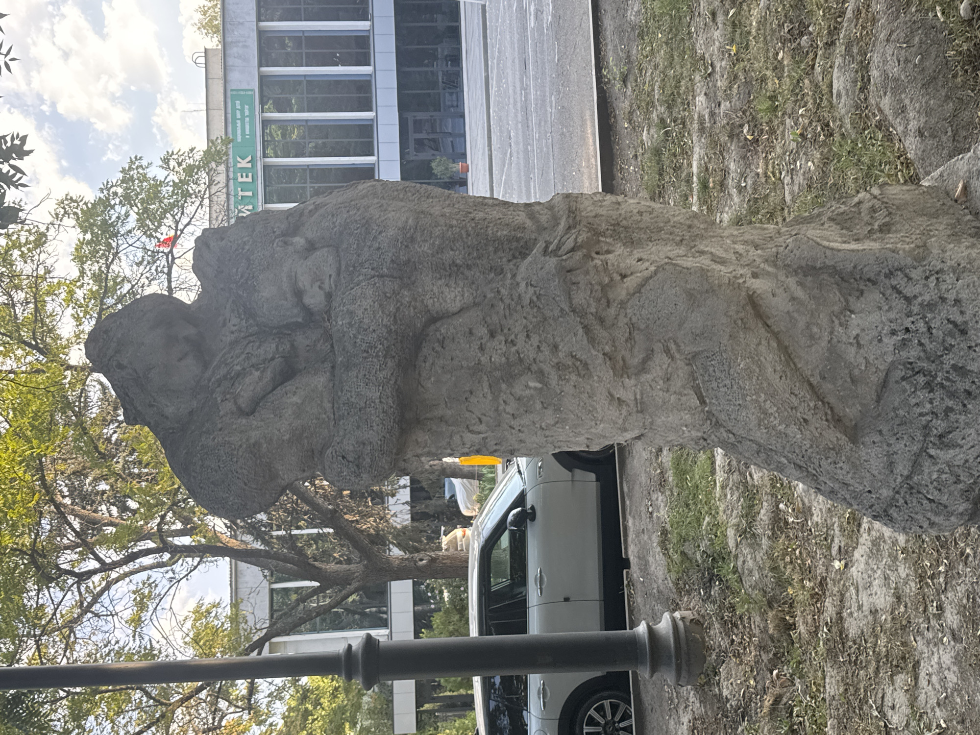 In Bishkek, Kyrgyzstan, a weathered stone sculpture depicts two figures embracing. The sculpture stands on a patch of dry grass and dirt, with fallen leaves scattered around its base. In the background, a white Mini Cooper is parked near a lamppost. Beyond the car, a modern building with a green sign that reads "АПТЕК" (Apteka, meaning pharmacy) is visible. Trees with green leaves and a cloudy sky complete the urban landscape.