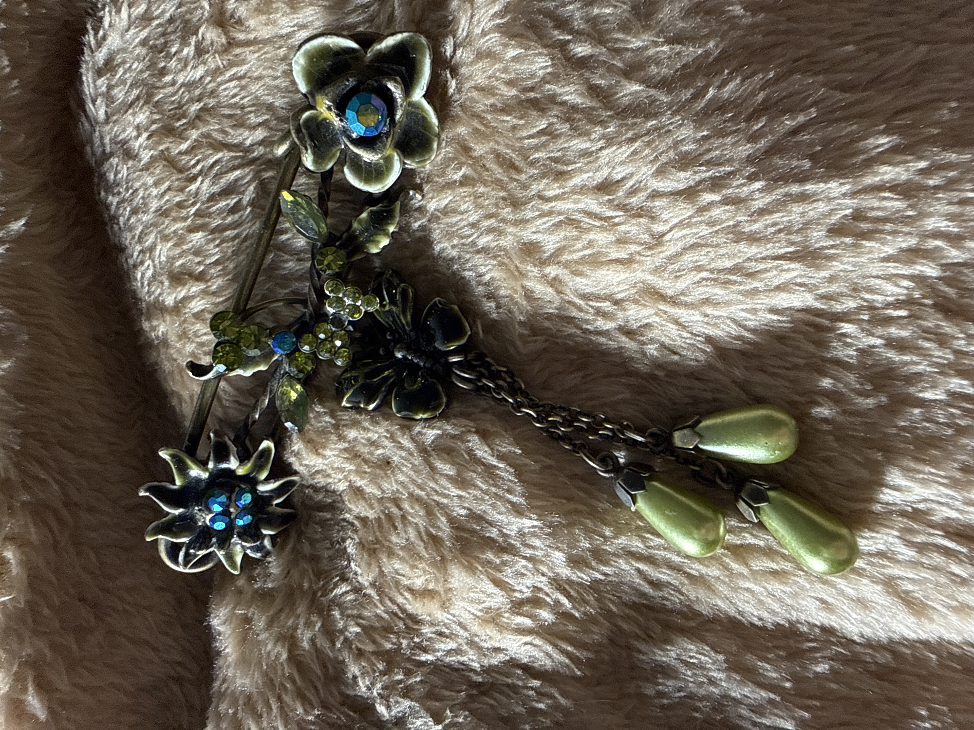A decorative pin is lying on a soft, brown, furry surface. The pin features two flower-like designs, connected by a stem with green leaves and small green and blue gemstones. The top flower has a larger, iridescent blue gemstone in the center. Three teardrop-shaped, light green pendants hang from the side of the pin via short, tarnished chains.