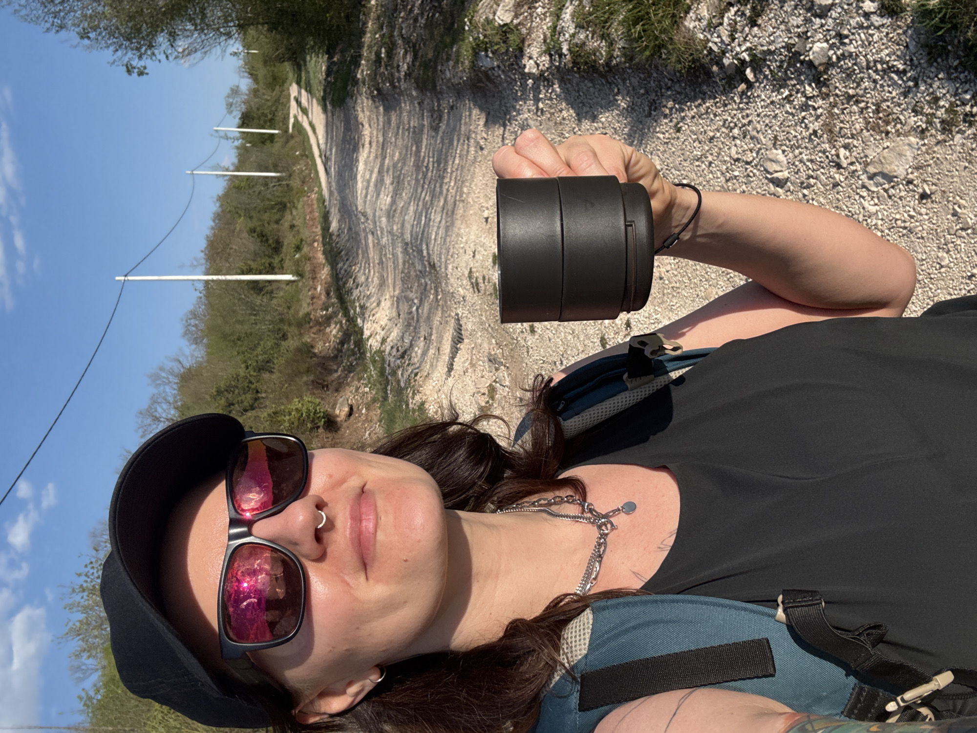 A selfie shows a person wearing sunglasses, a black baseball cap, a silver necklace, a black shirt, and a blue backpack, holding a black cylindrical object in their right hand. The person has a silver nose ring. The background reveals a dirt path surrounded by vegetation. White poles, likely for electrical or communication lines, are visible in the distance along the path. The scene appears to be outdoors in Podbadac, Montenegro.