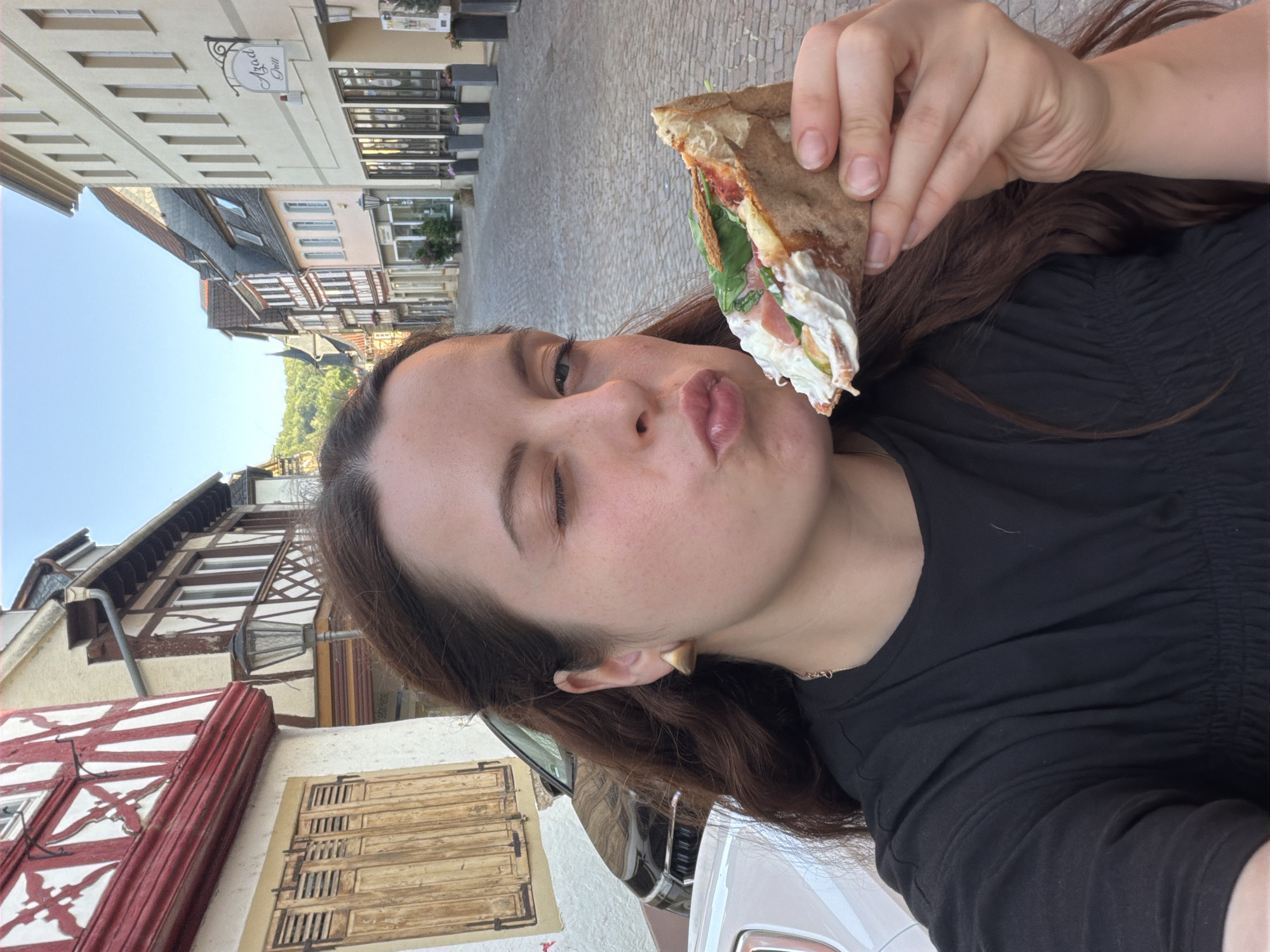 In Meisenheim, Germany, a woman with long, dark hair, wearing a black top and a gold earring, is captured in a close-up selfie as she winks and puckers her lips. She holds a slice of pizza adorned with various toppings including lettuce, tomato, and what appears to be ham, up to her mouth. The background reveals a charming European street scene with cobblestone pavement and traditional half-timbered buildings, characteristic of the town's historic architecture. A black car is parked on the left side of the street, and the facade of a building on the right displays a sign that reads "Azad Grill." The setting sun casts a warm glow over the scene.