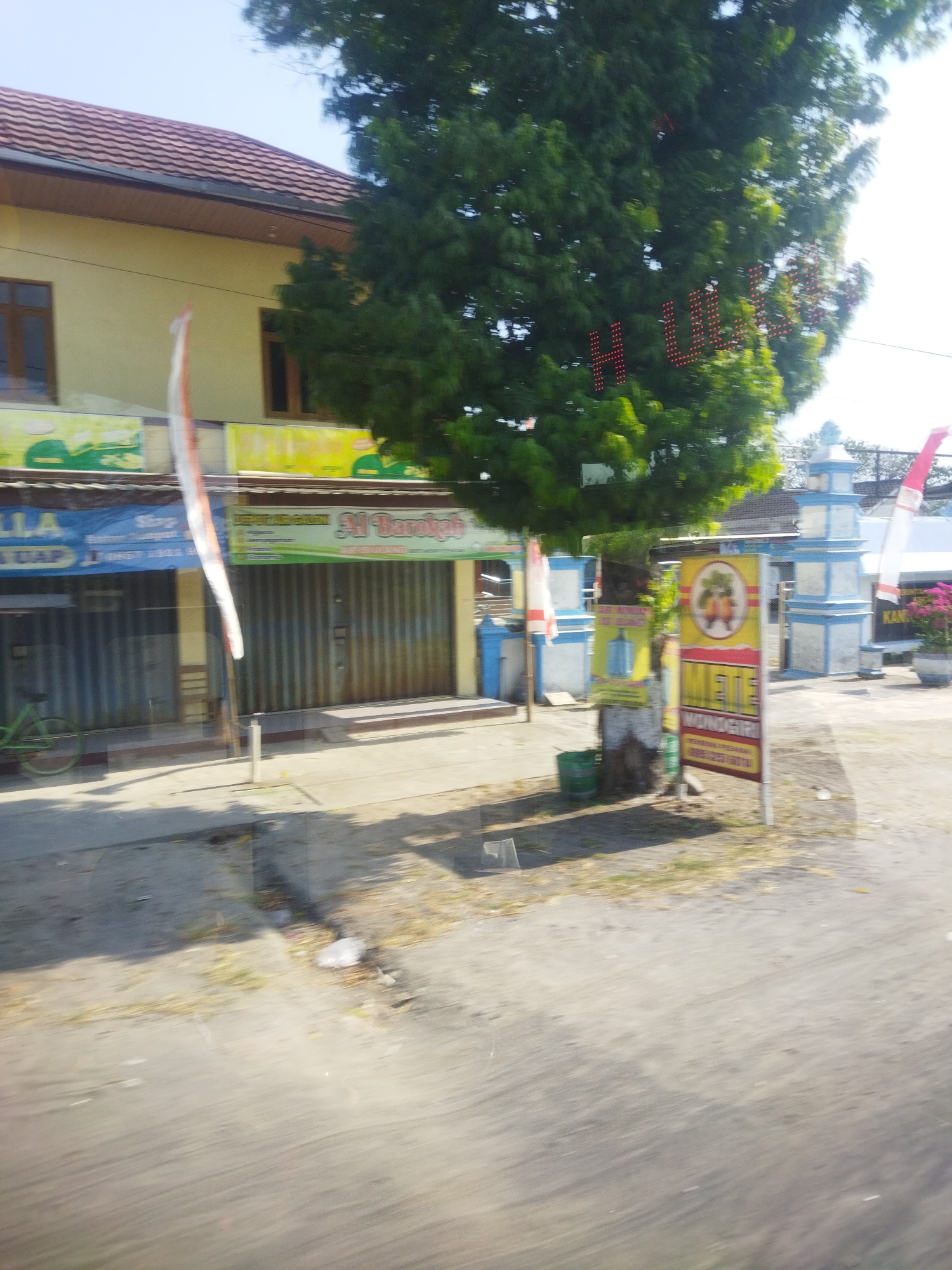 This is a low-angle, medium shot of a street scene in Nguter, Indonesia, taken on a sunny day. The foreground shows a paved road with some debris and shadows from trees. In the middle ground, there are several buildings with storefronts. The building on the left is yellow with a brown roof and windows. A white and red flag is attached to a pole in front of it. To its right, there are two storefronts with metal roll-up doors. One sign reads "Al Barokah." A large green tree with red lights spelling out "H... Ul" is prominent in the center of the image. To the right of the tree, there is a blue and white decorative structure, possibly a gate or archway, with a flag attached. A red and yellow sign with images of fruit and text is also visible near the tree. A green bicycle is parked in front of one of the storefronts. The background shows more buildings and vegetation under a clear blue sky.