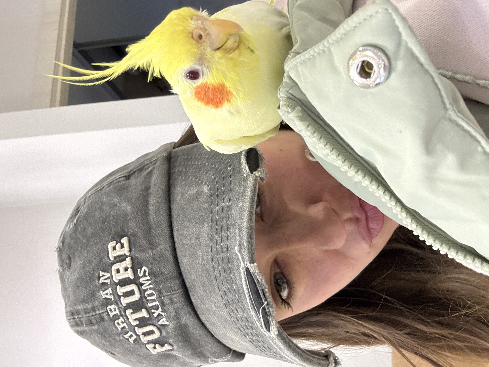 A woman is holding the camera at a close angle to her face. A yellow cockatiel is perched on her shoulder. The woman is wearing a grey baseball cap with the text "URBAN FUTURE AXIOMS" embroidered on it. She is wearing a light green jacket. The background is out of focus. The location is not discernible from the image.