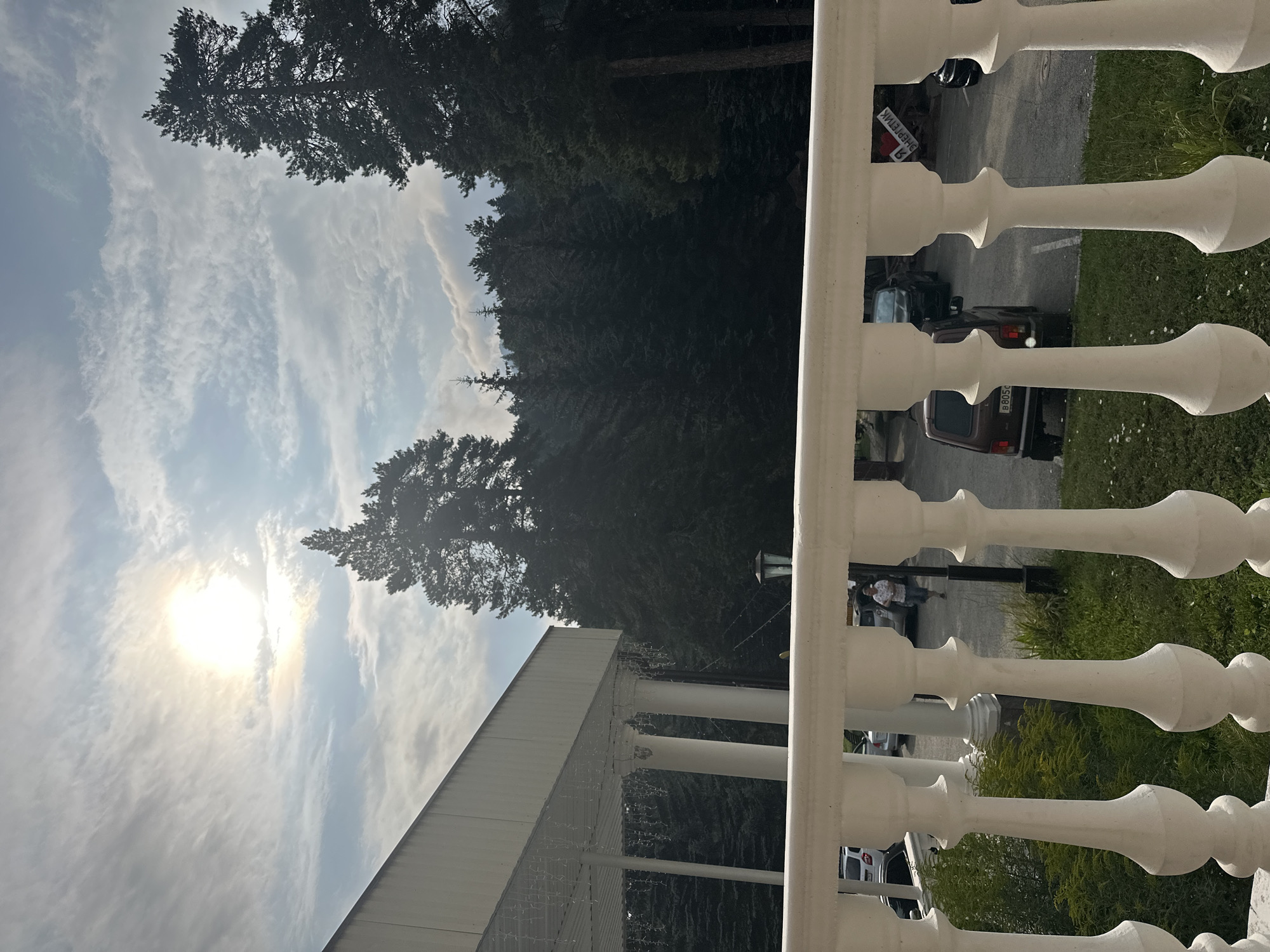 The image shows a bright, sunny day with a partly cloudy sky over a forested area. In the foreground, a white ornate balustrade is visible. Beyond the balustrade, there is a paved area that appears to be a parking lot, with several vehicles parked. A person is walking across the parking lot. In the background, tall evergreen trees form a dense forest. To the left, there is a white building with columns, suggesting it might be a hotel or a resort. The overall scene is one of a natural setting, possibly a tourist destination, in a place like Arkhyz, Russia, known for its mountainous terrain and natural beauty.