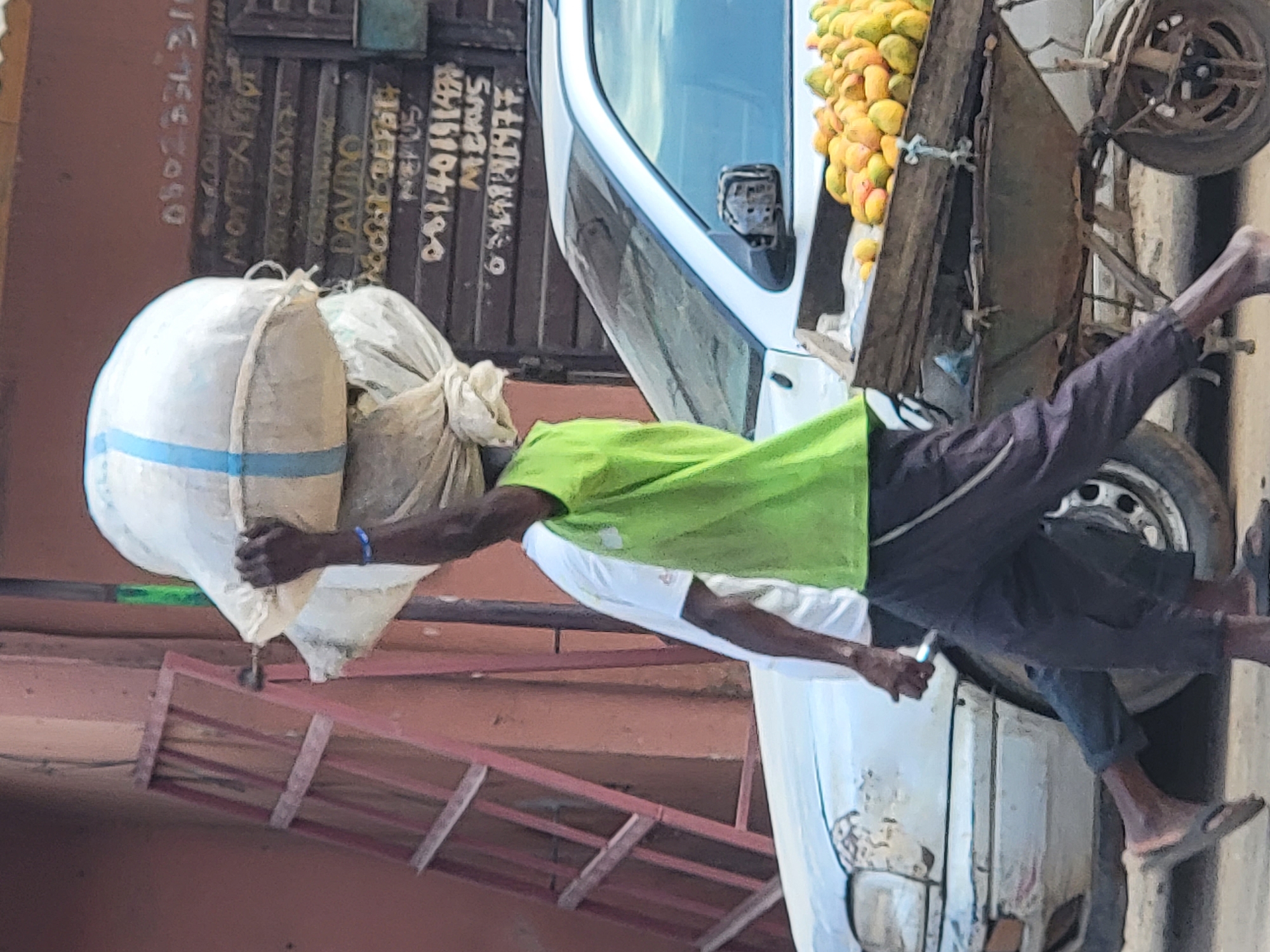 In Maiburiji, Nigeria, a man wearing a green shirt and dark pants rides a three-wheeled cart filled with yellow fruit. He is seated behind the driver, whose back is to the viewer and who is wearing a white shirt and dark pants. They are transporting two large sacks, one with a blue stripe, secured to the side of a building with numbers painted on it. A red ladder rests against the same building.