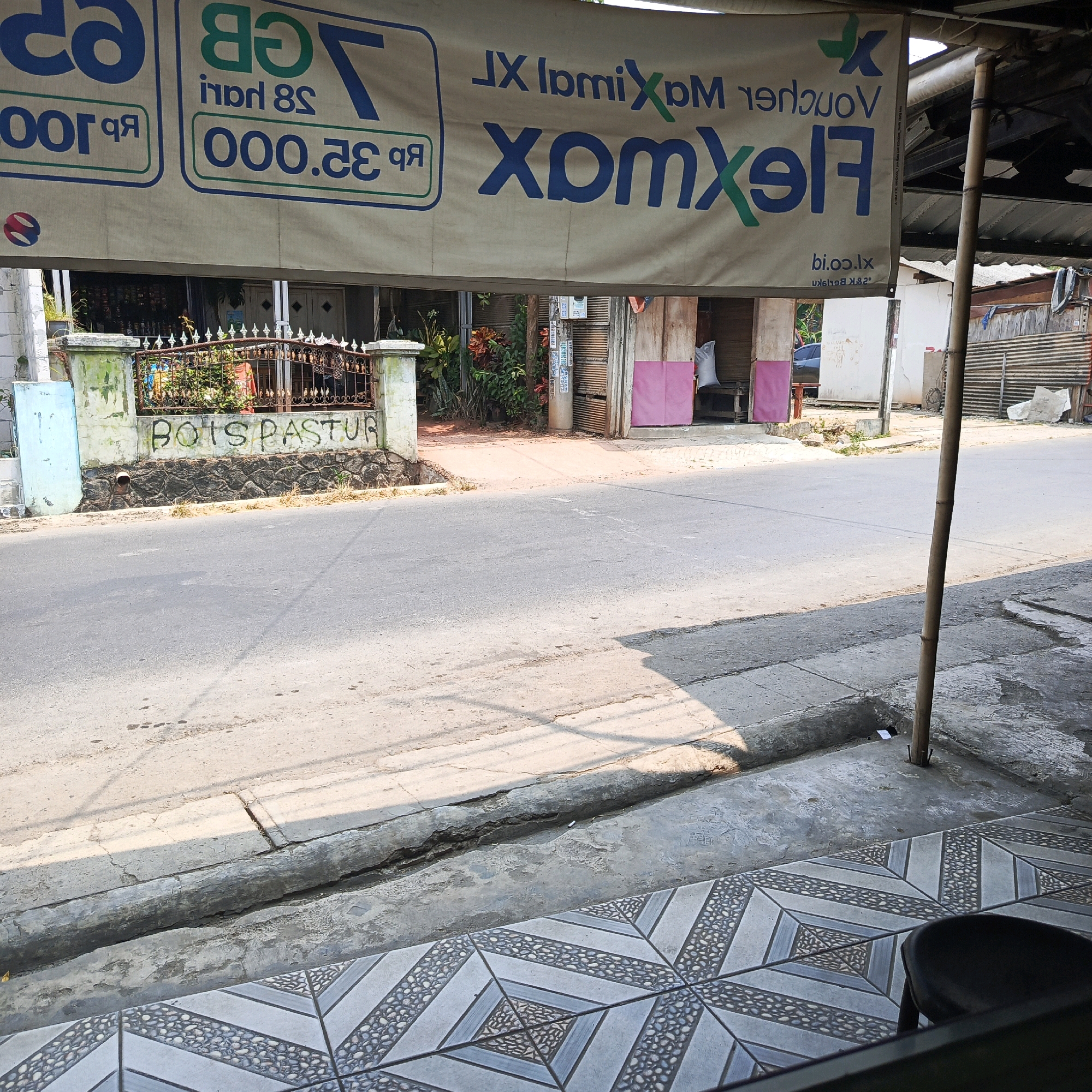 The image shows a street scene in Karawang, Indonesia. A banner advertising "VOUCHER MAXI" is displayed prominently. Below the banner, graffiti on a wall reads "BOIS PASTRU". The street is paved, and there are buildings and vegetation visible in the background. A bamboo pole stands vertically on the right side of the frame. The overall scene suggests a commercial area with a mix of advertisements and street art.
