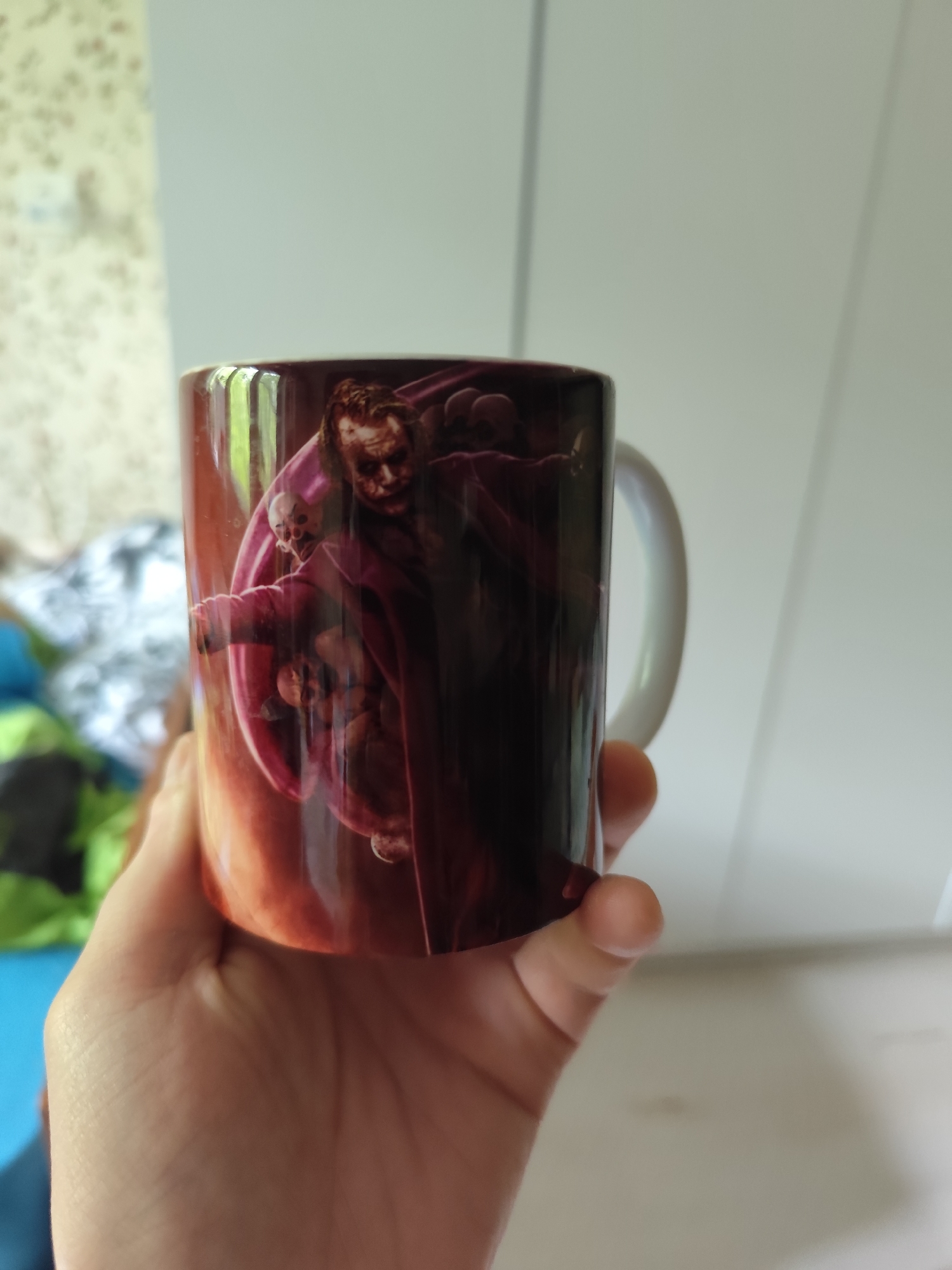 A person's hand holds a mug indoors in Moscow, Russia. The mug features a vivid illustration of the Joker character, identifiable as Heath Ledger's portrayal from "The Dark Knight," dressed in a dark purple coat with a sinister grin. Around the central Joker figure, several smaller, skull-like or skeletal figures are depicted. The artwork on the mug is dark and dramatic, with a gradient background of reddish-brown and orange hues.
The background of the image is softly blurred, indicating a shallow depth of field. To the right, a large, plain light-grey or white surface, possibly a wall or a closet door with vertical seams, dominates the backdrop. To the left, a portion of a patterned wall or fabric with light, floral-like designs is visible. Below that, some brightly colored, out-of-focus objects in green and blue suggest clothes or household items.
The lighting is bright and appears to be natural, suggesting it is daytime. No specific activity beyond holding the mug is taking place, and no text is visible on the mug or in the background.