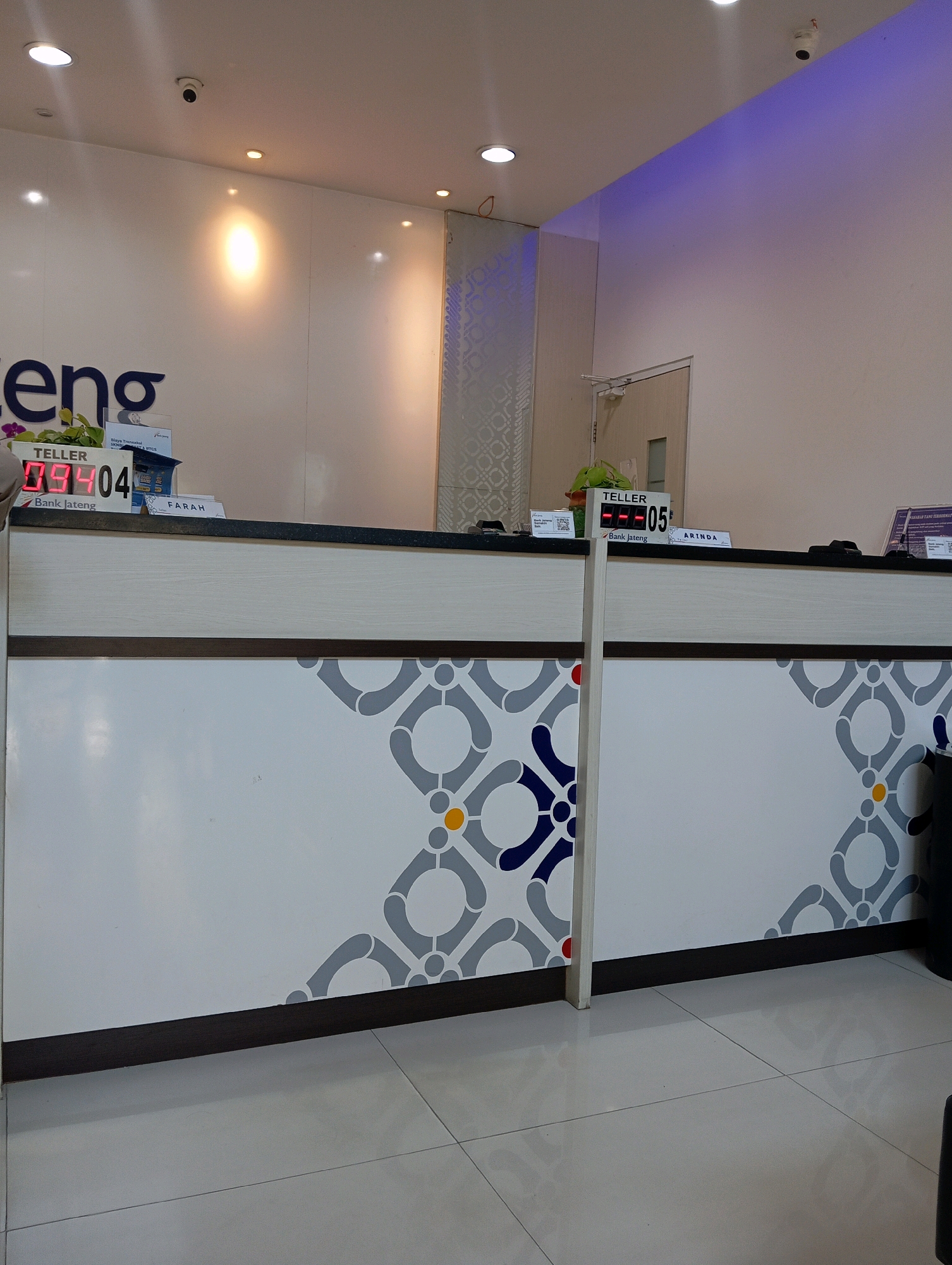 The image shows the interior of a bank lobby in Magelang, Indonesia. The scene is focused on a teller counter with digital displays indicating customer queue numbers. On the left, a digital display reads "TELLER 09404" above "Bank Jateng". On the right, another display reads "TELLER 05" with "Bank Jateng" below it, and a nameplate "ARINDA" is visible. The counter is decorated with a grey, white, and blue geometric pattern. In the background, a door and a section of wall with a patterned texture are visible. Lighting fixtures are present on the ceiling, and a security camera is mounted high on the wall. The floor is tiled in a light grey color. No people are clearly visible in the frame, but the setup suggests active bank operations.