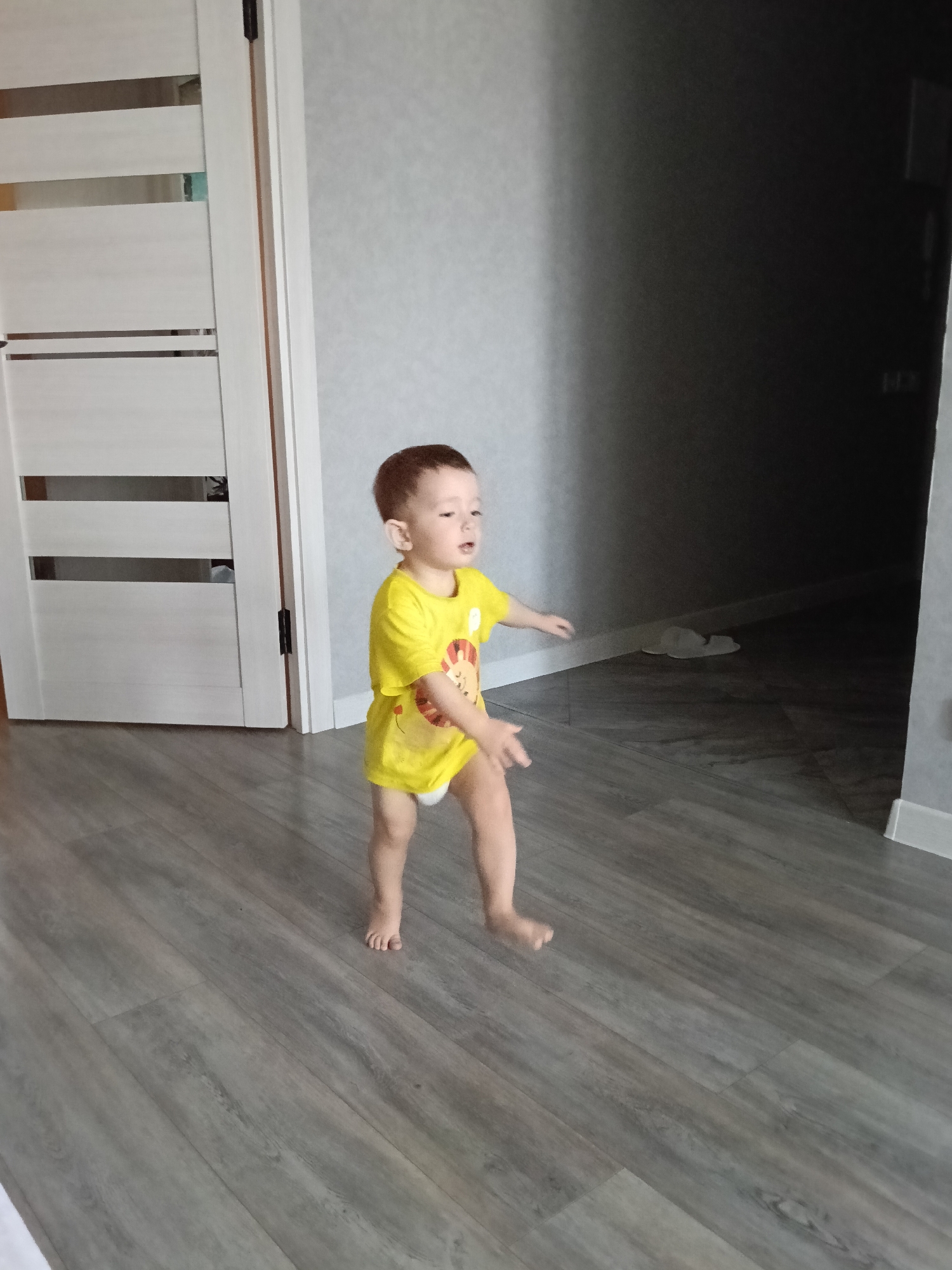 The photograph depicts a young child, likely a toddler, walking barefoot on a gray, wood-grain patterned floor inside a residence. The child is wearing a yellow t-shirt with a cartoon lion on it, and appears to be wearing a diaper. A white interior door with horizontal decorative panels is visible to the left of the child. The room transitions into a darker area in the background. The location is determined to be Murom, Russia, based on available metadata or contextual information not explicitly provided in the image.