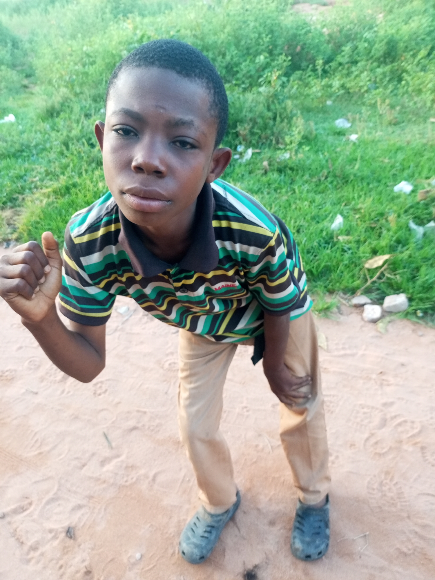 A young boy with dark skin and short, dark hair poses outdoors, looking towards the camera. He is wearing a striped polo shirt with black, green, yellow, and white horizontal stripes, and tan trousers. He is standing on a sandy surface with green foliage and some scattered white plastic debris in the background. His left hand is raised and slightly clenched. His right hand is near his right knee. He is wearing dark blue clogs on his feet. The scene appears to be in a rural or semi-rural area, potentially in or near Tempure, Nigeria.