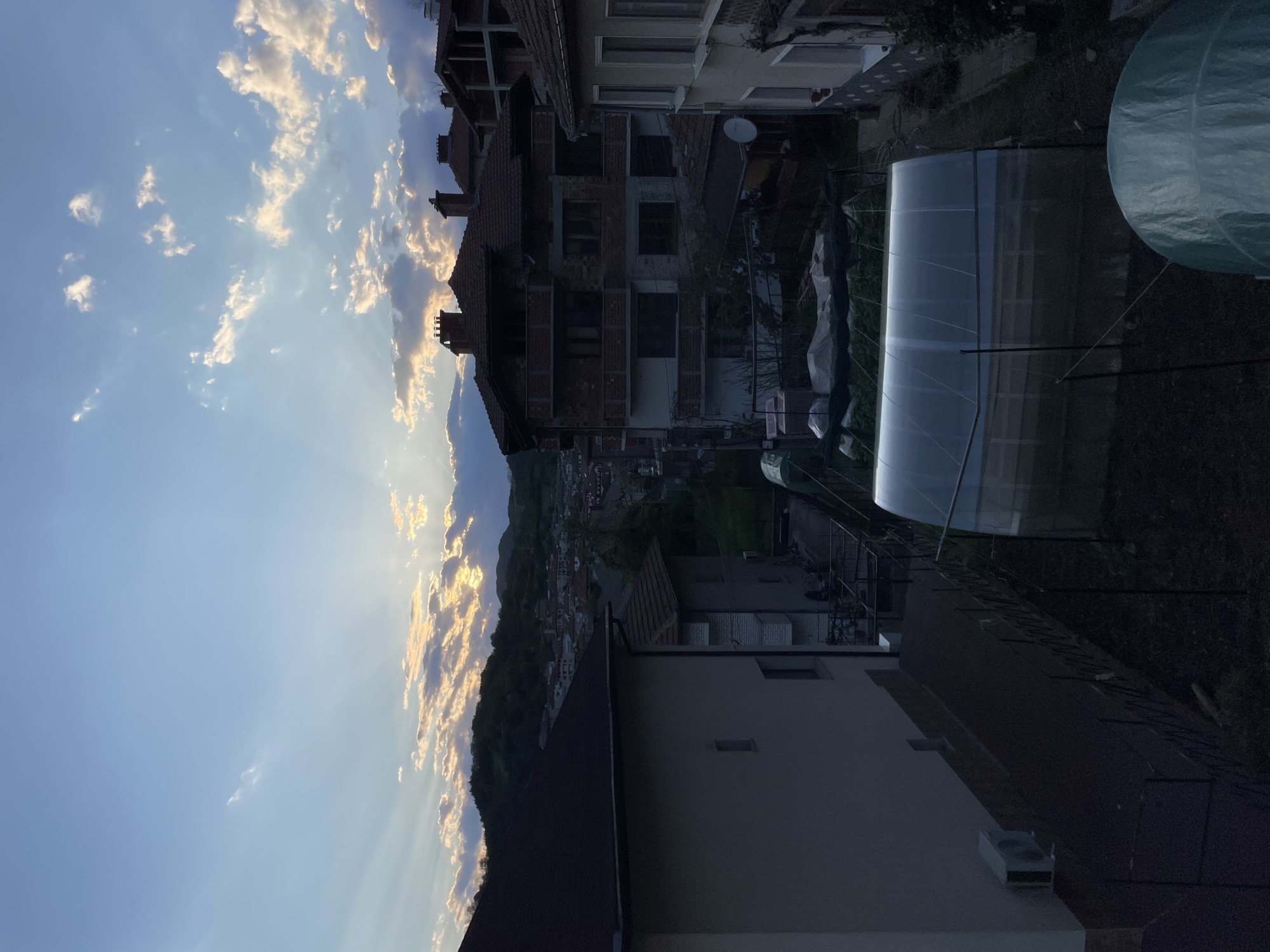 A rotated, outdoor shot overlooking Zlatograd, Bulgaria, shows a residential area at dusk. A clear, blue sky with scattered clouds stretches across the upper left, with rays of sunlight breaking through. A forested mountain range is visible in the middle ground. Houses with red-tiled roofs and various architectural styles line the landscape. A greenhouse with a translucent plastic cover sits in a garden, partially visible in the dark right side of the image.