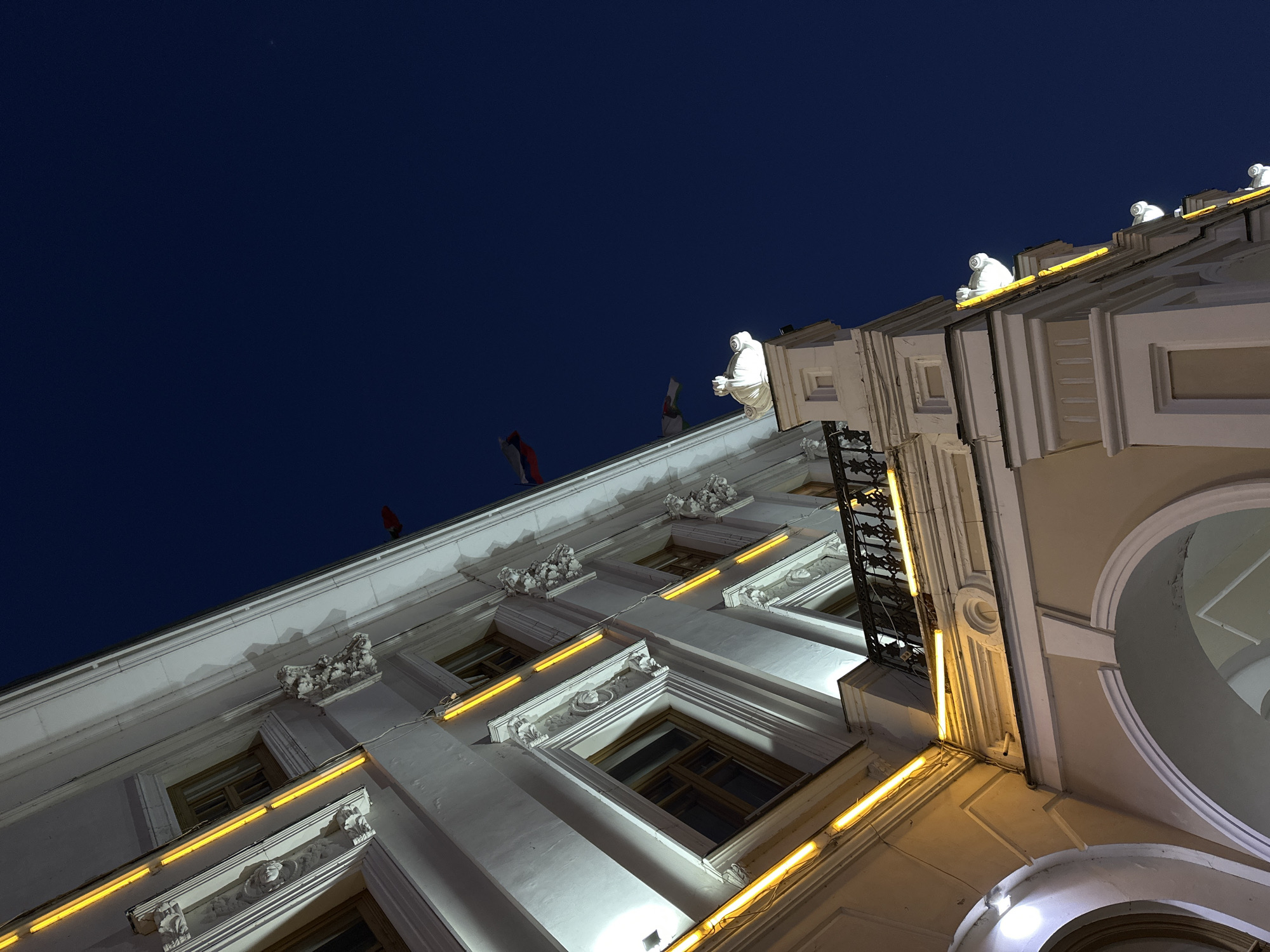 An upward-facing shot of the facade of a light-colored building in Kazan, Russia, taken at night. The building's architectural features are prominent, including multiple windows with decorative trim and moldings. Small sculptures adorn the roofline, illuminated by a light source. Two small flagpoles with red flags on top are mounted to the roof. A dark blue sky fills the background. String lights are placed along the building.