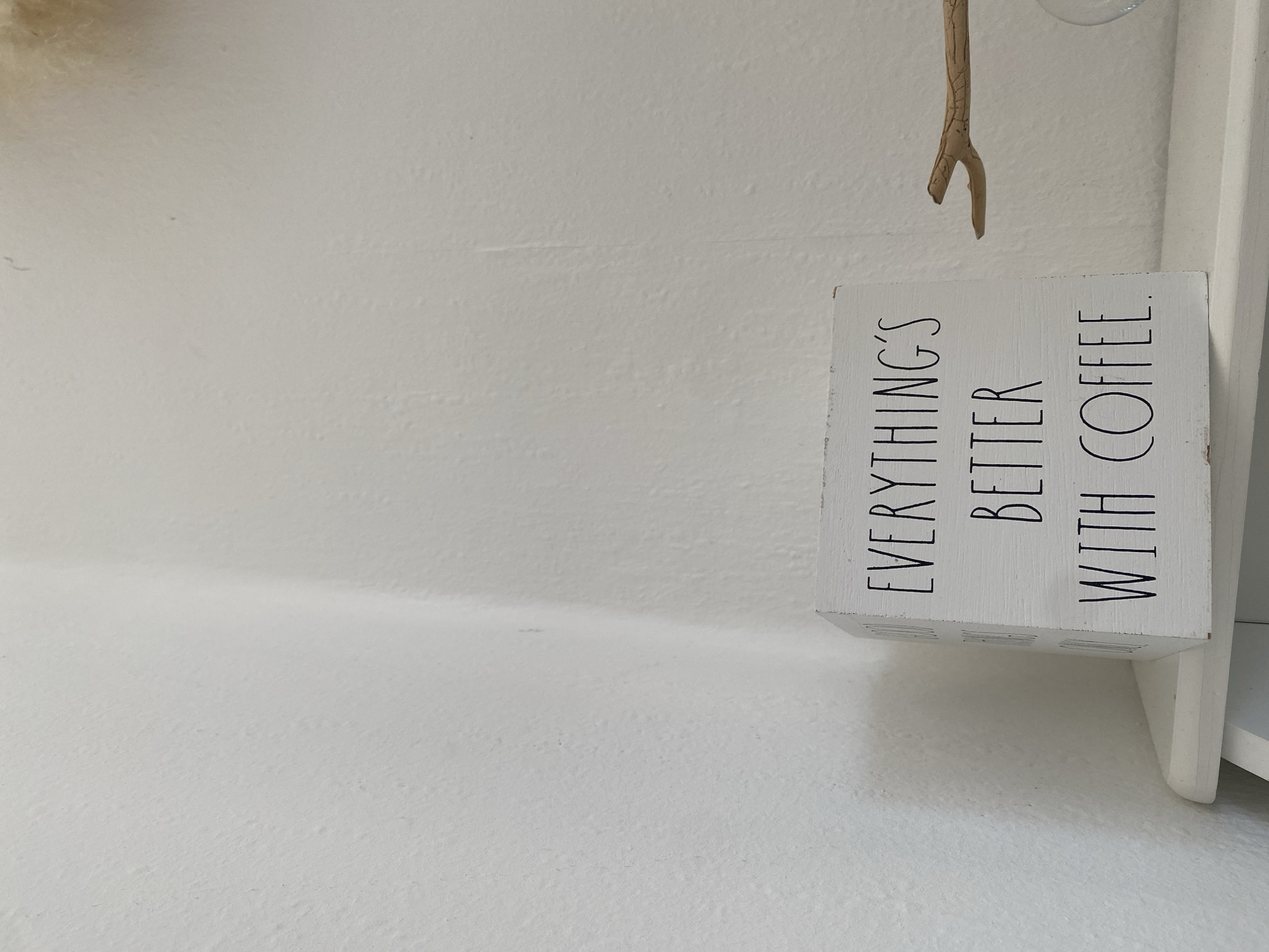 A white wooden cube with black text sits on a white shelf. The text reads, "EVERYTHING'S BETTER WITH COFFEE." To the right of the cube, a small twig rests on the shelf, pointing towards a glass object. The background is a white wall with subtle texture and a visible vertical seam. This image was captured in Bad Sachsa, Germany.