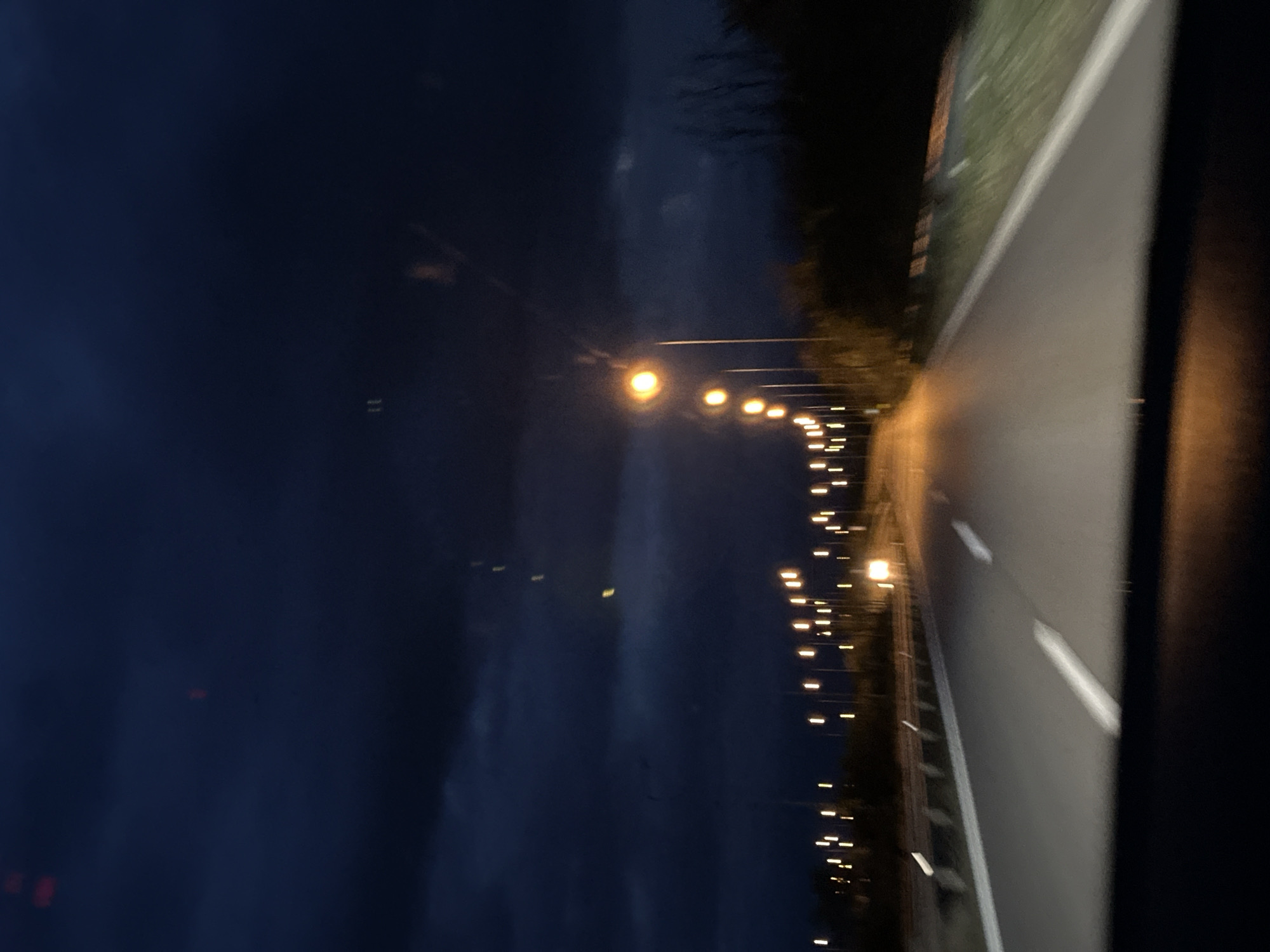 A nighttime view along a highway illuminated by streetlights, taken from inside a vehicle. A row of lights stretches into the distance along the road's edge on the right. The highway has a white line marking the lanes. The sky is dark, and some trees are visible on the right. The exact location in Κάτω Χριστός, Greece, cannot be determined without additional visual or contextual information.
