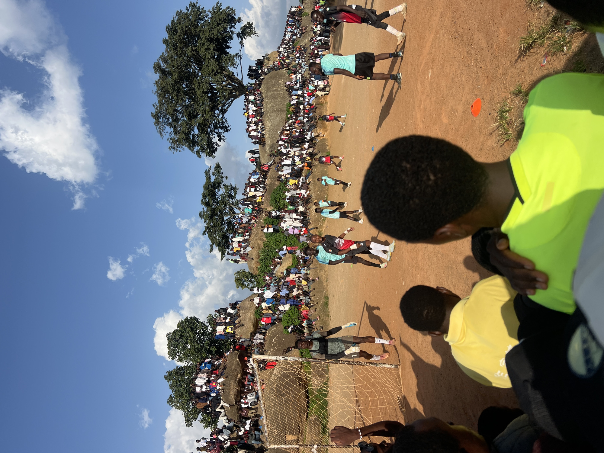 The image depicts an outdoor soccer game in Jos, Nigeria. The field is made of dirt, and a goal with a net is visible on the left side of the frame. Numerous spectators are gathered on a rocky hillside overlooking the field, creating a vibrant and crowded atmosphere. The sky is blue with scattered white clouds. Several players are on the field, actively engaged in the game. In the foreground, the backs of several spectators are visible, suggesting the photo was taken from within the crowd. The overall scene captures a lively community event with a strong sense of local engagement.
