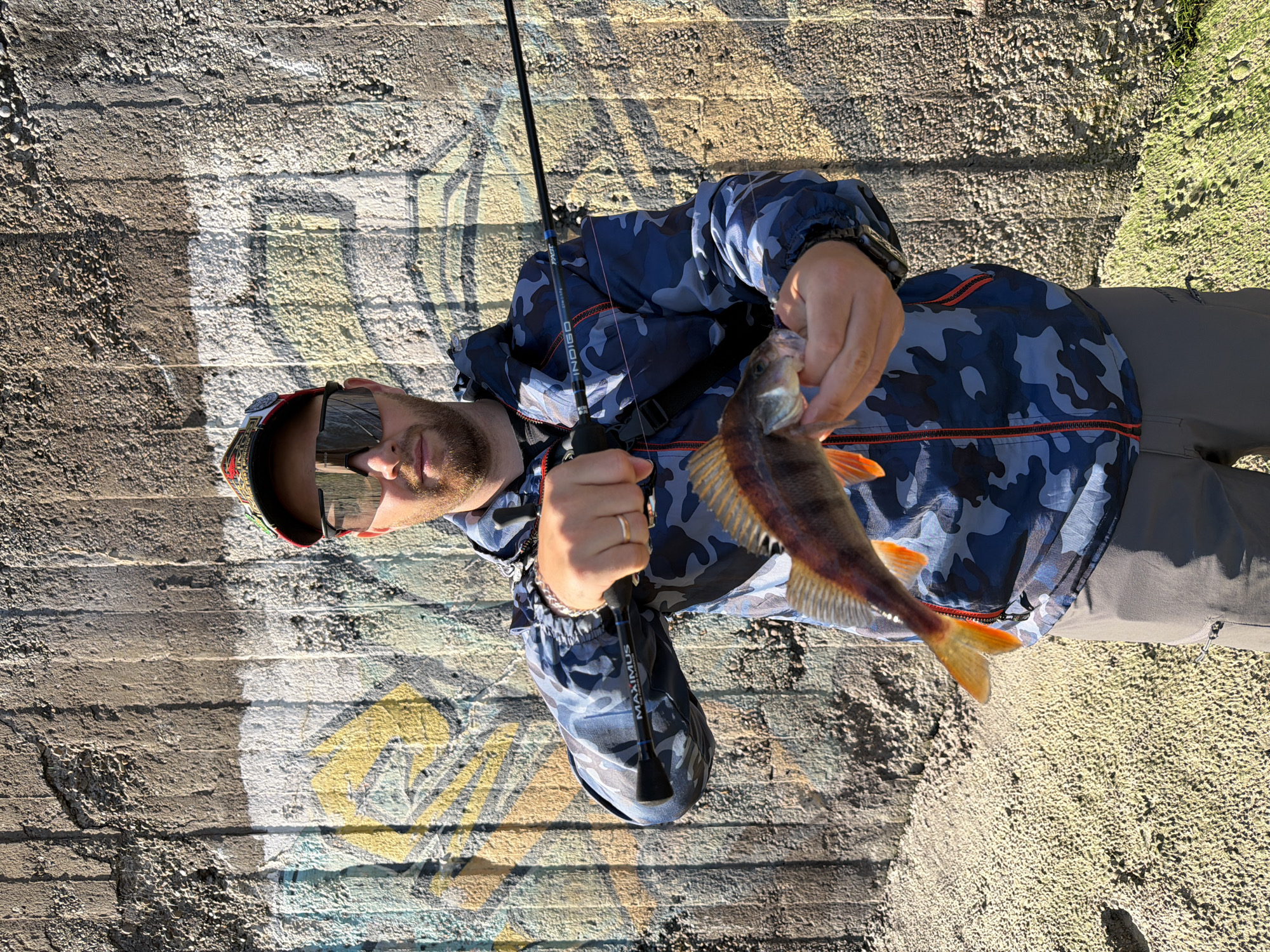 A man is standing on a concrete surface with graffiti and holding a fish in his right hand. He is also holding a fishing rod in his left hand. He is wearing sunglasses, a hat, a blue camouflage jacket, gray pants, and a watch. The location is likely Chaikovsky, Russia, as implied by the user's request. The background consists of a weathered concrete structure covered with graffiti and green vegetation. The scene suggests the man has been fishing.
