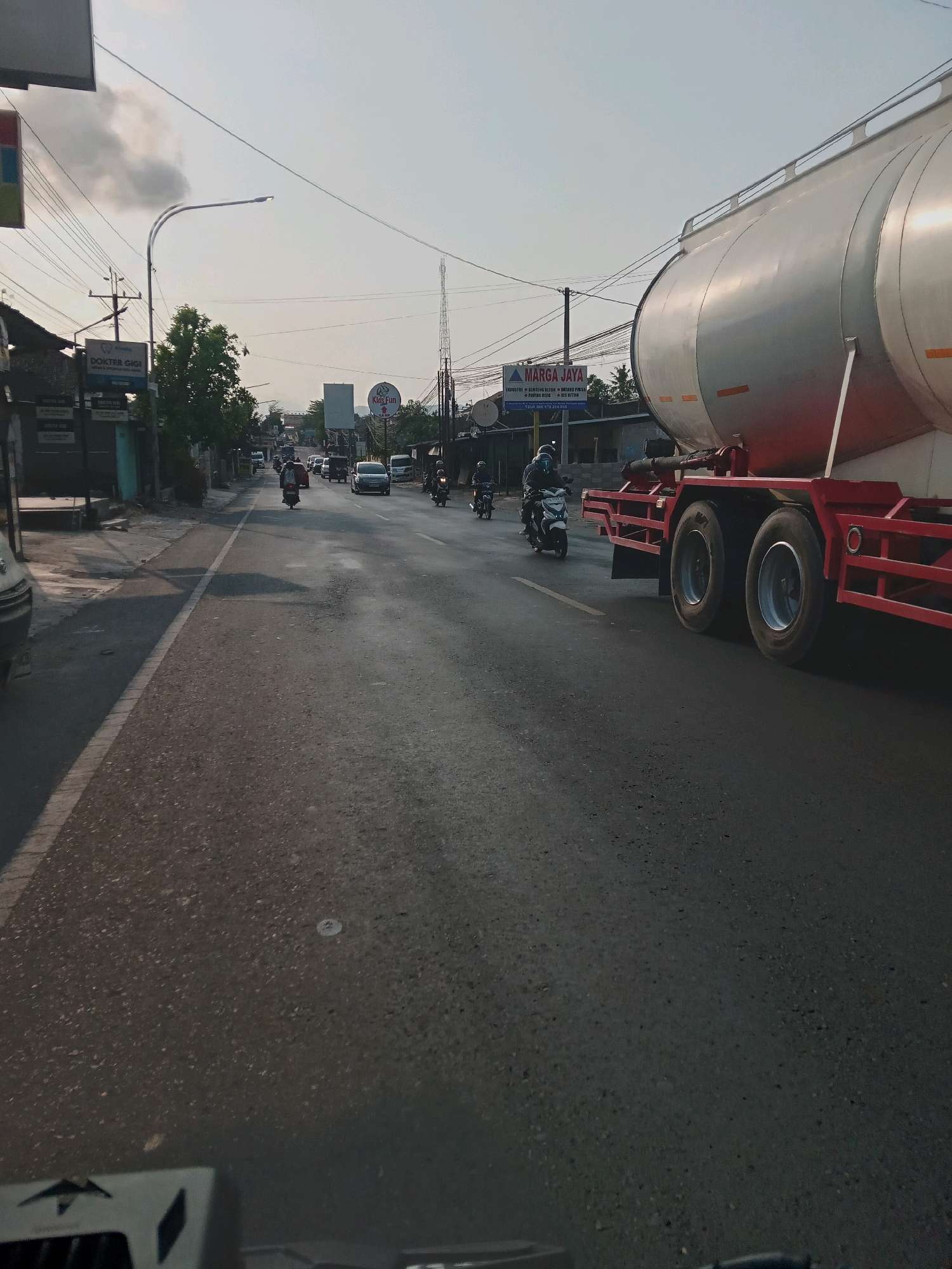 The image shows a street scene in Berbah, Indonesia, captured from the perspective of a vehicle, likely a motorcycle, given the partial view of handlebars and a dashboard-like object in the foreground. The scene is illuminated by natural daylight, possibly late afternoon, with a hazy sky.
The road is paved and appears to be a two-lane street with a white line dividing the lanes. Traffic is present, with several motorcycles and at least one car visible. In the foreground, a large tanker truck is prominently positioned on the right side of the frame, its red chassis and silver tank being the dominant elements. The truck is moving, as indicated by its position on the road.
On the left side of the road, there are buildings, including signs that appear to be for businesses. One sign reads "DOKTER GIGI," indicating a dentist's office. Further down the street, there are trees and utility poles with overhead wires. A tall communication tower is visible in the distance. Another sign on the right side reads "MARGA JAYA," with additional text below, suggesting a business or company. A circular sign with "Kids Fun" is also visible.
The overall impression is of a busy street in a semi-urban or suburban area in Indonesia, with typical road infrastructure and commercial activity. The low angle of the sun creates shadows on the road and buildings.