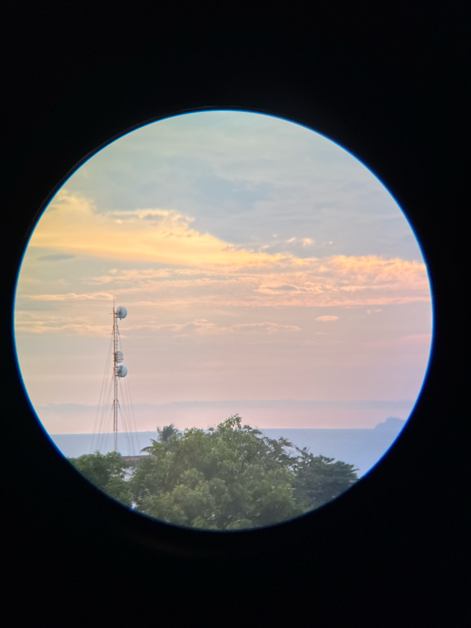 The image is a photograph taken through a circular lens, possibly a telescope or camera lens, looking out at a landscape at dusk. The scene is framed by a dark, blurred outer edge, with the view centered on a telecommunications tower. The tower is tall and metallic, with three large spherical antennas mounted at different heights.
Below the tower, lush green trees form a dense foliage line. Beyond the trees and the tower, a body of water, likely the sea, is visible, extending to the horizon. In the distance, a faint, mountainous island or landmass can be seen on the right side of the frame.
The sky above is filled with soft, pastel-colored clouds, suggesting either sunrise or sunset. The clouds are tinged with hues of orange, yellow, pink, and light blue, creating a serene and picturesque atmosphere.
The location in the city of Nha Trang, Vietnam is indicated by the presence of the telecommunications tower, which is a common sight in coastal cities, and the tropical foliage visible in the foreground. The overall impression is one of a peaceful twilight view overlooking the sea and the city's infrastructure.