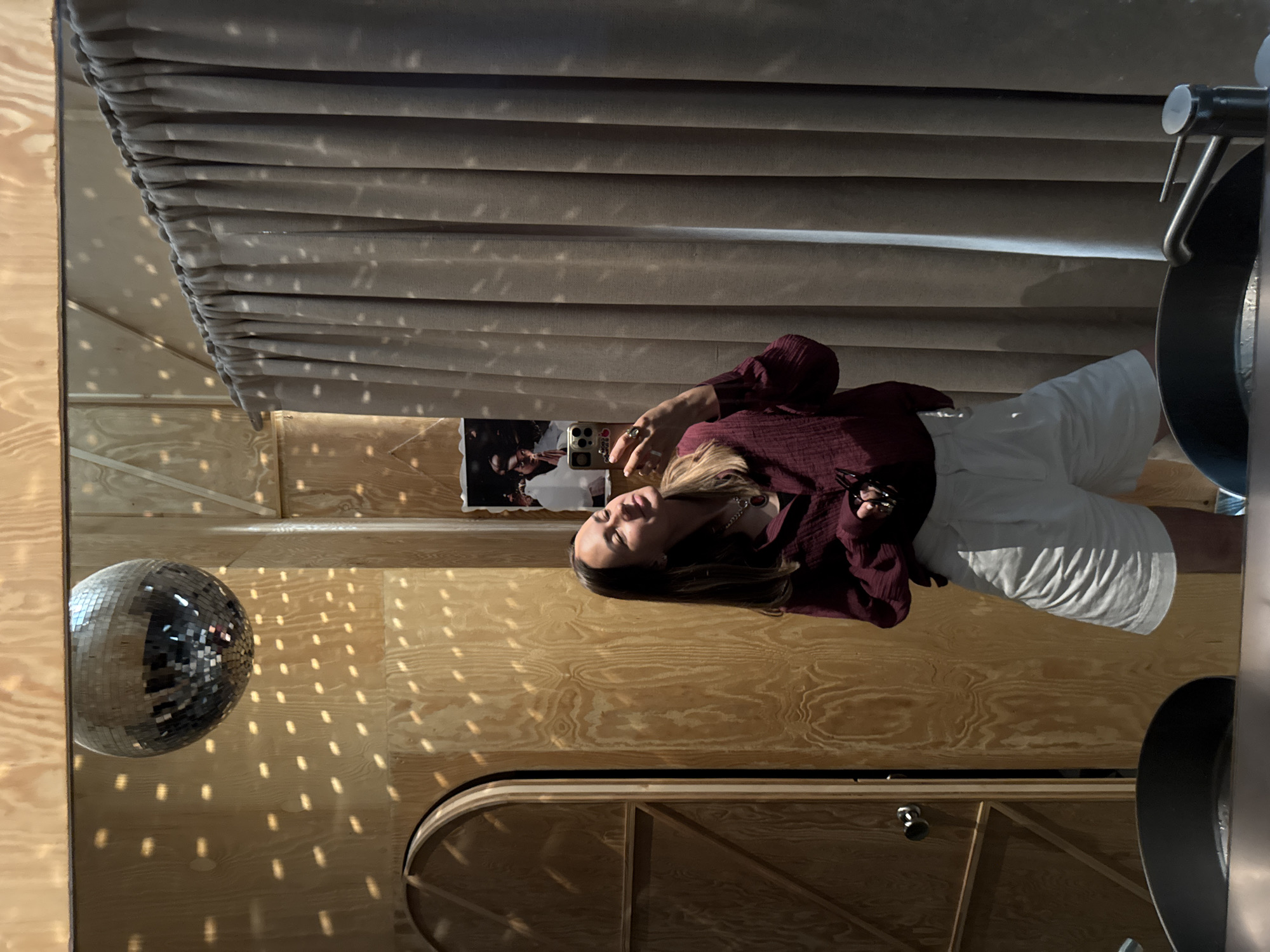 The image shows a young woman taking a selfie in a room with wooden walls and a disco ball. She is wearing a burgundy shirt and white shorts. The room appears to be a bathroom, as there is a sink and a faucet visible in the foreground. The disco ball is hanging from the ceiling and casting reflections on the walls. There is a large curtain on the right side of the image, and a door on the left. The overall ambiance is dimly lit and somewhat retro.
