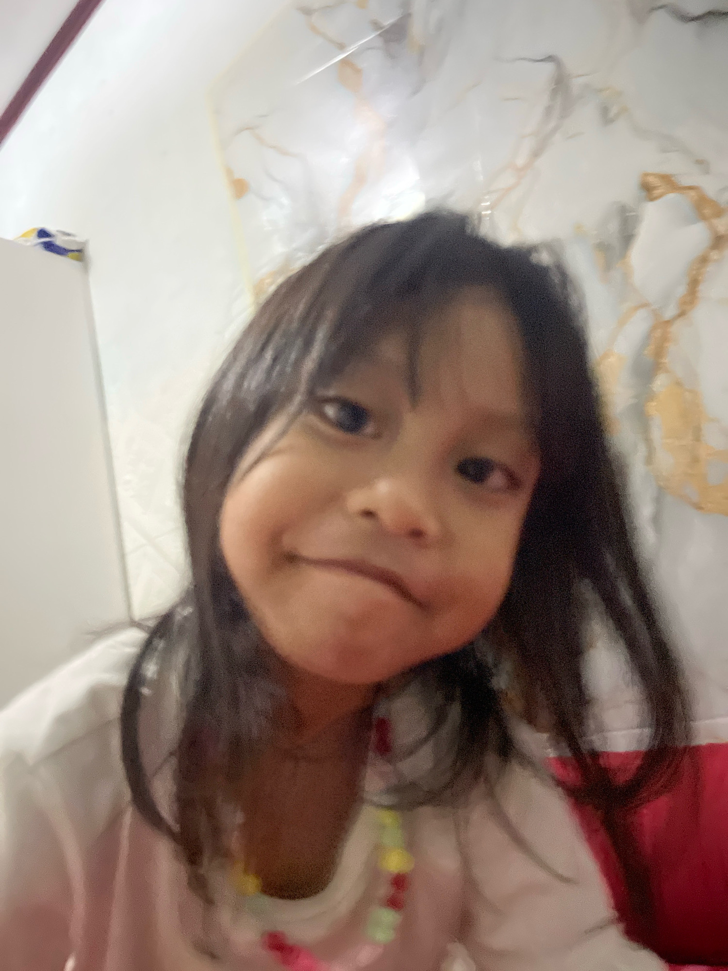 A close-up, blurry, selfie-style photograph shows a young East Asian child with dark hair and dark eyes. The child is looking directly at the camera with a slight smile. The child is wearing a light-colored shirt, possibly pink and white, with a colorful beaded necklace. The background is out of focus but appears to be a wall with a marble-like pattern, in a domestic setting. The photograph is taken in the city of Kecamatan Pati, Indonesia.