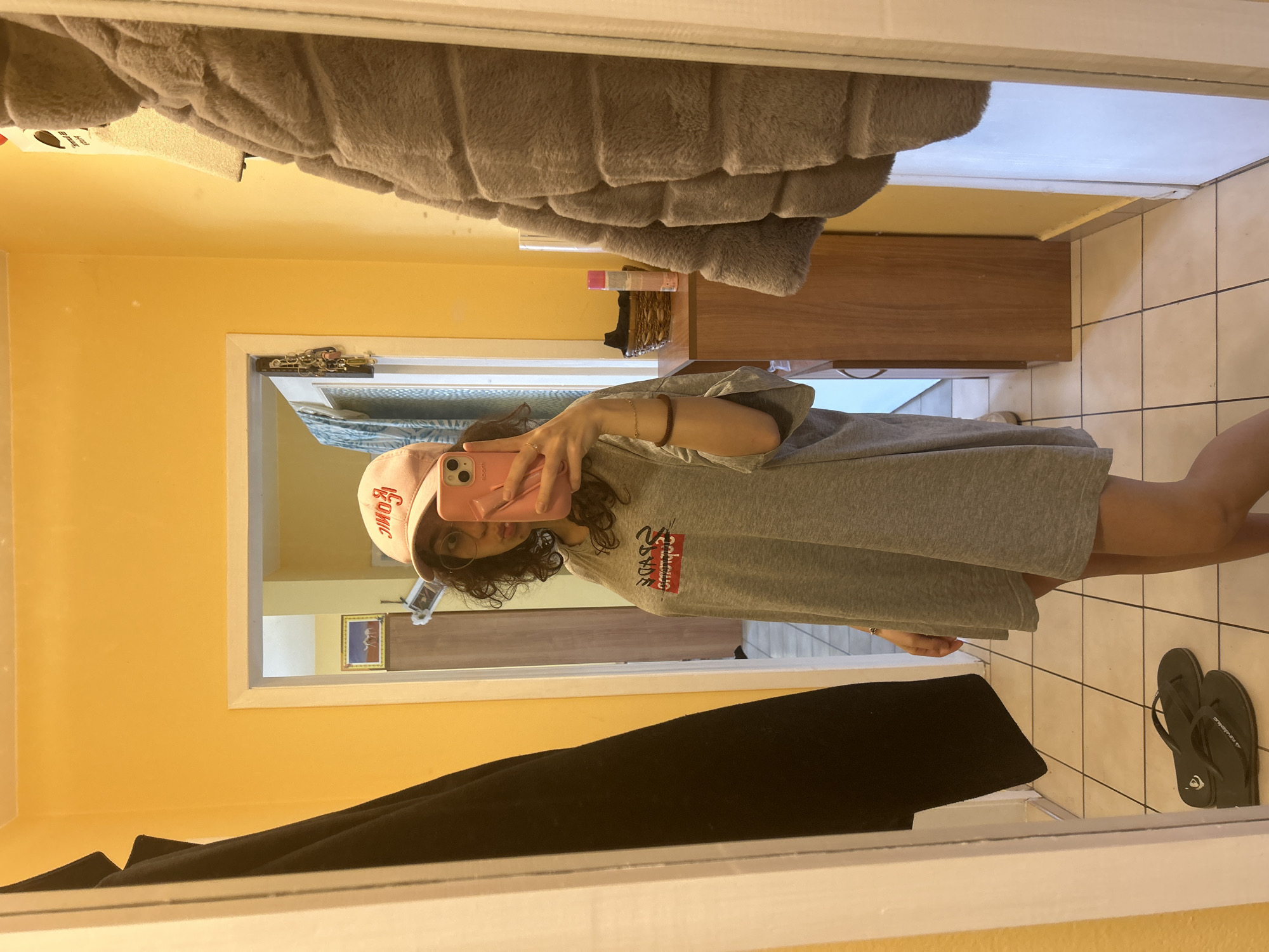 A person is taking a mirror selfie in a room with yellow walls. The person is wearing a grey t-shirt and a pink hat. There are sandals on the tiled floor. A bed is visible above, and a dark-colored garment hangs in the lower part of the frame. A wooden cabinet is visible in the background. The specific location in Sofia, Bulgaria, cannot be determined from this image.
