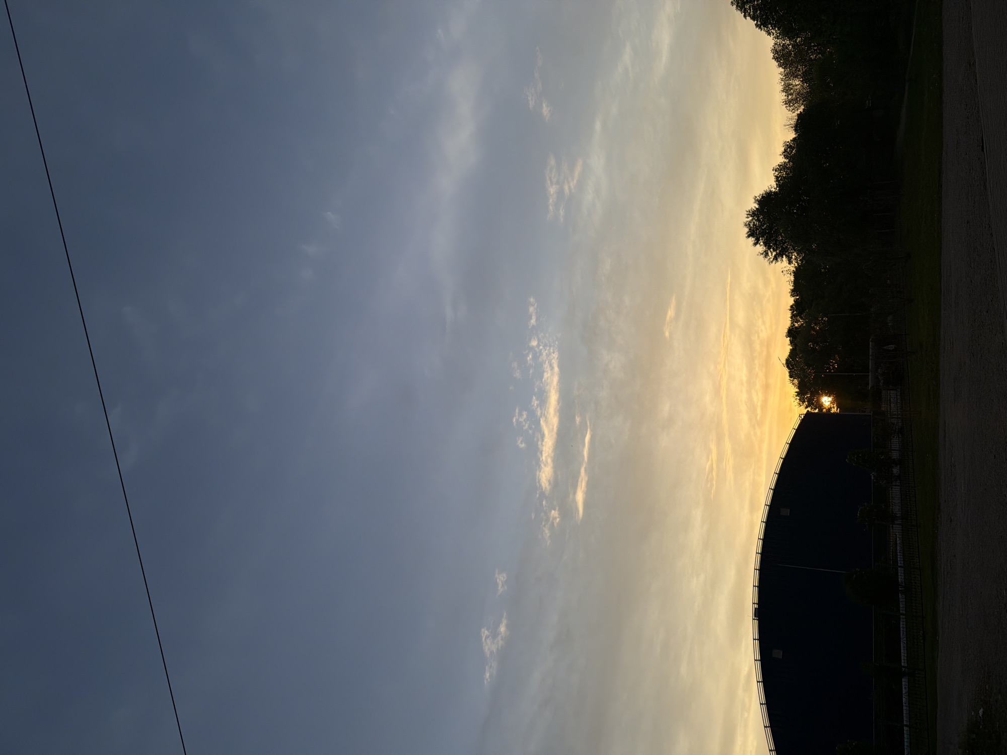The image shows a sky with the sun setting, casting a golden glow on the horizon. The left side of the image displays a blueish-gray sky with some clouds. In the lower right of the image, there is the upper curve of a large, dark building. The sun is visible through the trees and between the gaps of the building. A power line extends diagonally through the sky on the left. The location of Birobidzhan, Russia is not determinable from the provided visual information.