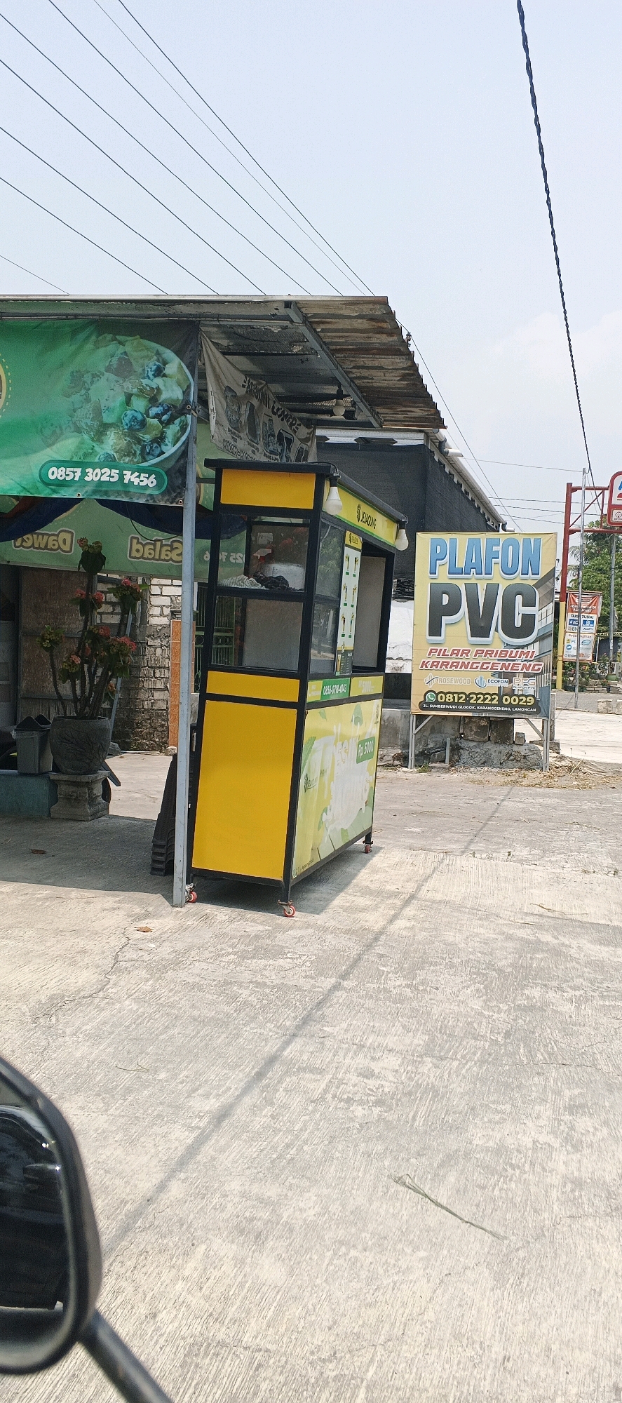 In a scene observed from the perspective of a vehicle, likely a motorcycle given the visible mirror in the foreground, a street-side vendor stall stands prominently. The stall is a yellow and black kiosk, designed for quick service, with glass panels showcasing its contents. Adjacent to it, a green banner displays food items and a phone number, "0857 3025 7456". Another banner, predominantly blue, advertises "PLAFON PVC" and provides contact information including a phone number "0812-2222 0029" and an address in "SUMBERWUDI GLOGOK, KARANGGENENG, LAMONGAN". The ground is paved with concrete, showing some cracks and debris. Overhead, power lines crisscross the frame against a pale sky. The environment appears to be an urban or semi-urban area in Indonesia, specifically referencing the village of Sumberwudi within the Karanggeneng subdistrict of Lamongan Regency. There are no people directly visible in the image, only the infrastructure and signage associated with commerce.