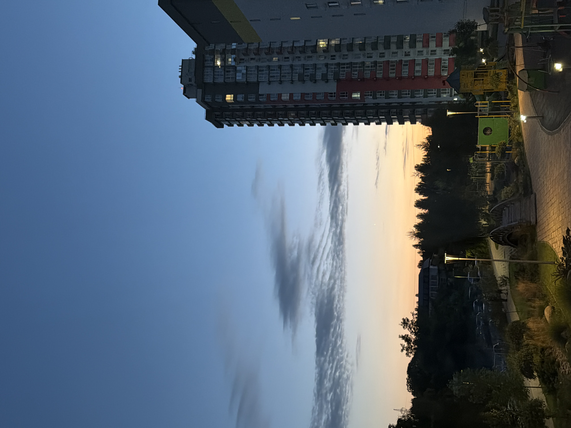 The image depicts an urban residential area in Kyiv, Ukraine, at dusk. A tall, modern apartment building with a red, white, and gray facade dominates the right side of the frame. Adjacent to it is another building, partially visible, with a gray exterior and yellow accents. The sky above is a gradient of deep blue, transitioning to lighter shades with wispy clouds and the soft glow of sunset on the horizon.
In the foreground, a well-maintained park area is visible. Paved pathways wind through landscaped grounds with trees, shrubs, and decorative rocks. Several modern lampposts illuminate the path. A children's playground with swings, slides, and climbing structures, some brightly colored in green and yellow, is present. A decorative arched bridge made of wood spans a small section of the park. In the background, a line of trees separates the park from a visible parking area with several cars. A building, appearing to be residential or possibly a community center, is situated on a hill behind the trees. The overall scene suggests a peaceful evening in a contemporary urban environment. No people are distinctly visible in the image.